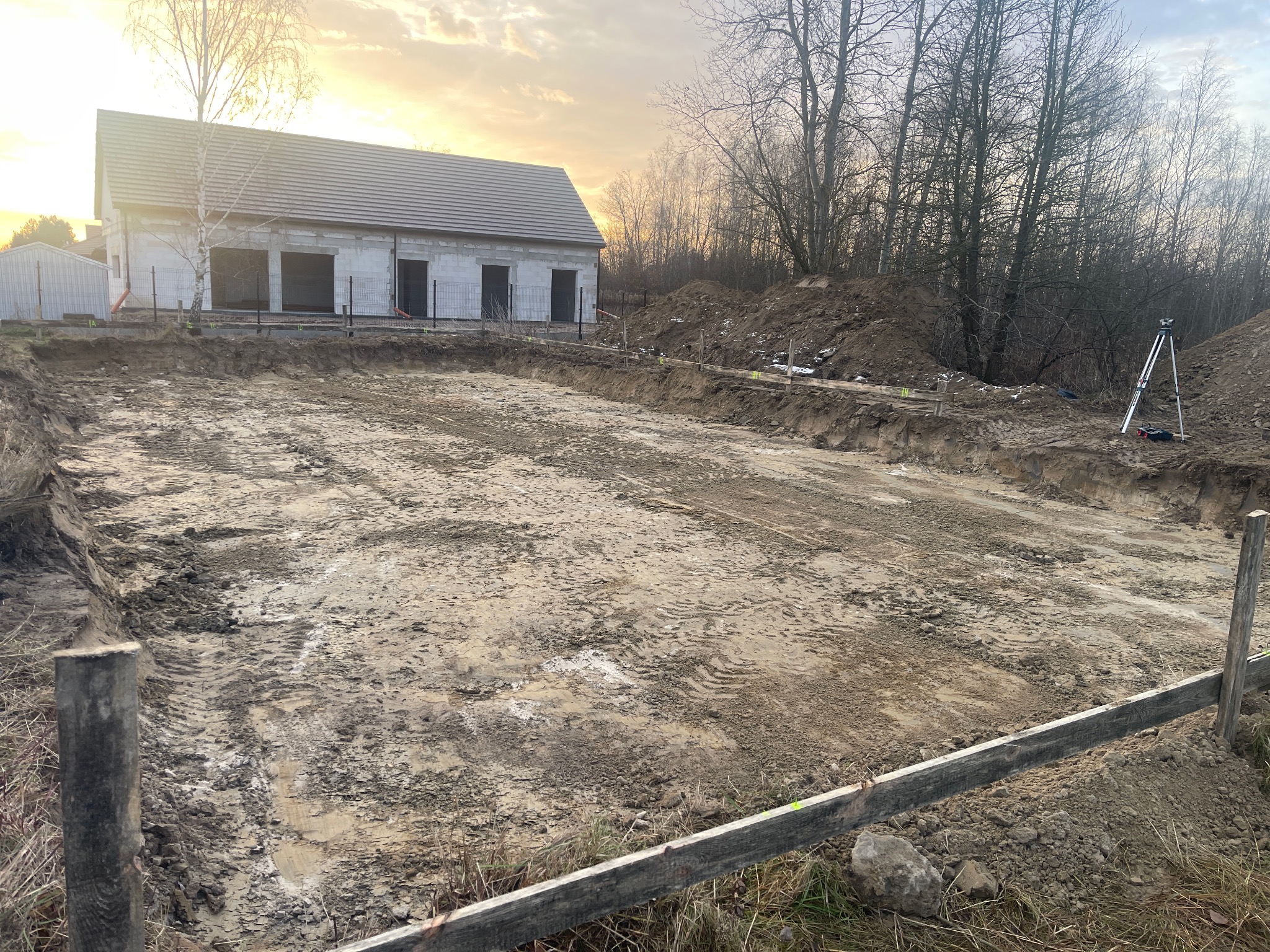 Wytyczony teren pod budowę domu jednorodzinnego z widocznymi fundamentami i częściowo wzniesionymi ścianami. W tle drzewa i zachodzące słońce.