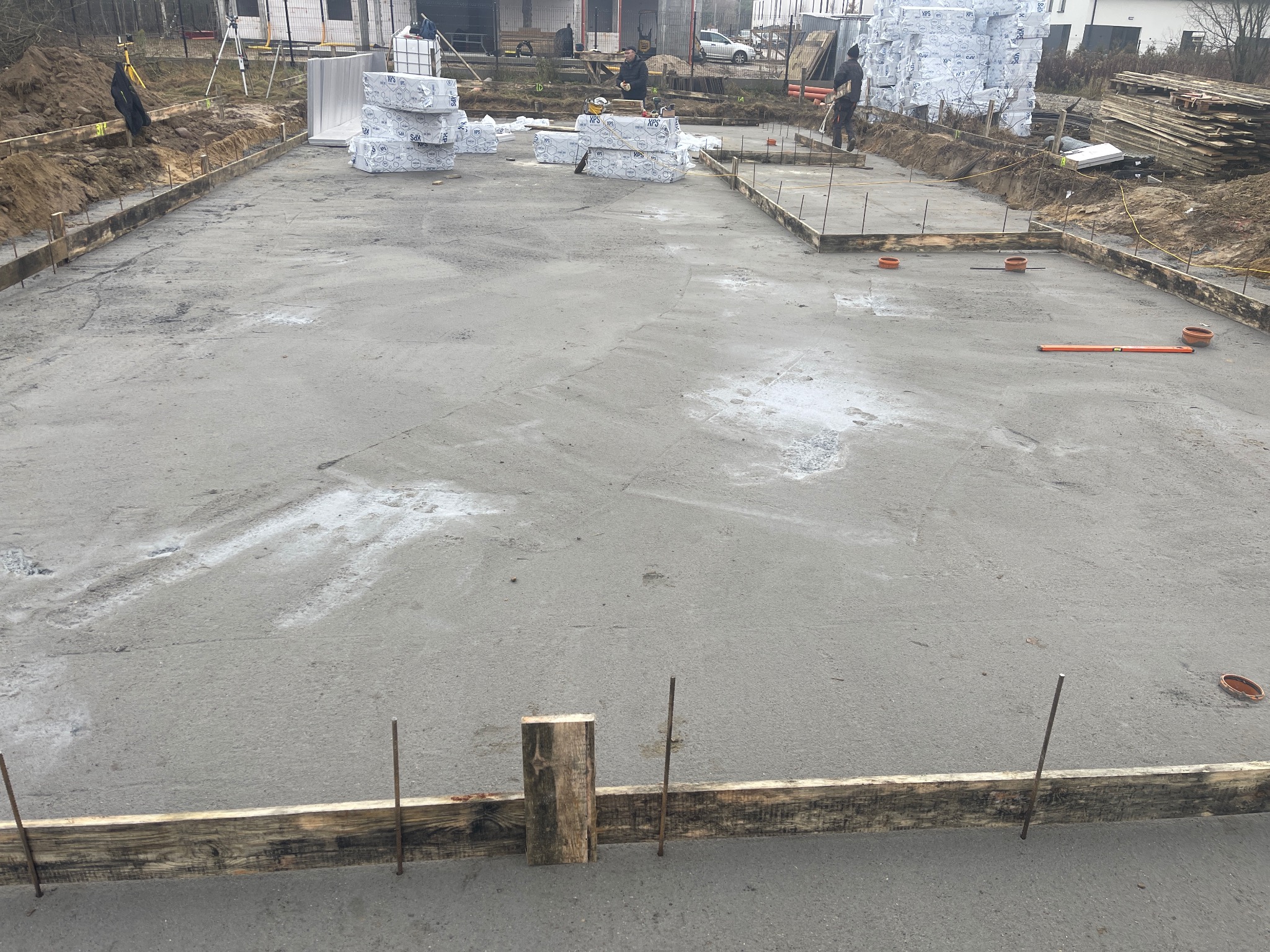 Wylana płyta fundamentowa z drewnianym szalunkiem i wystającymi prętami zbrojeniowymi. W tle materiały izolacyjne i pracownicy budowlani.