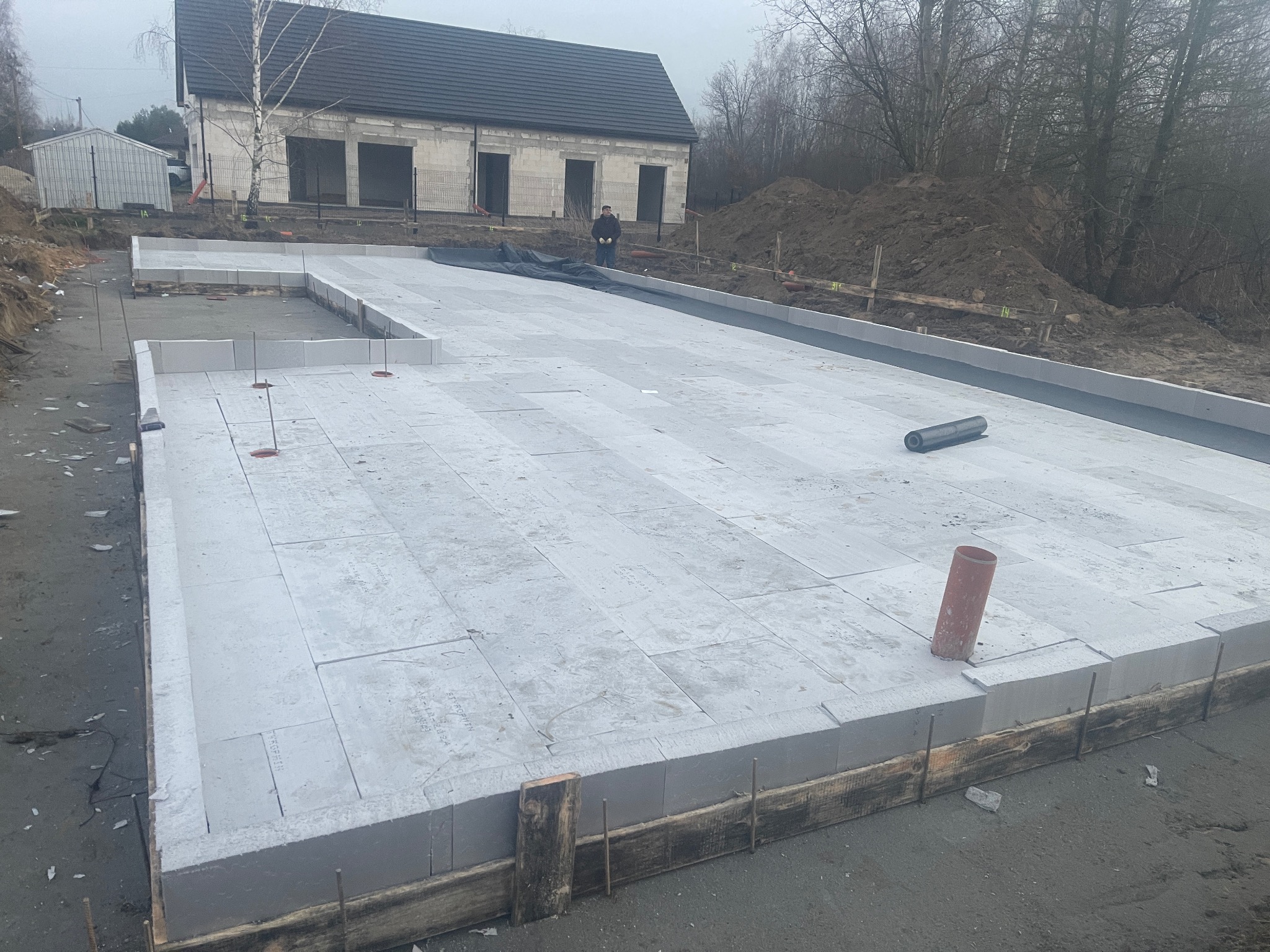 Fundament z bloczków betonowych na budowie domu jednorodzinnego, widok z góry. W tle surowy budynek i hałda ziemi. Na pierwszym planie wystające pręty zbrojeniowe.