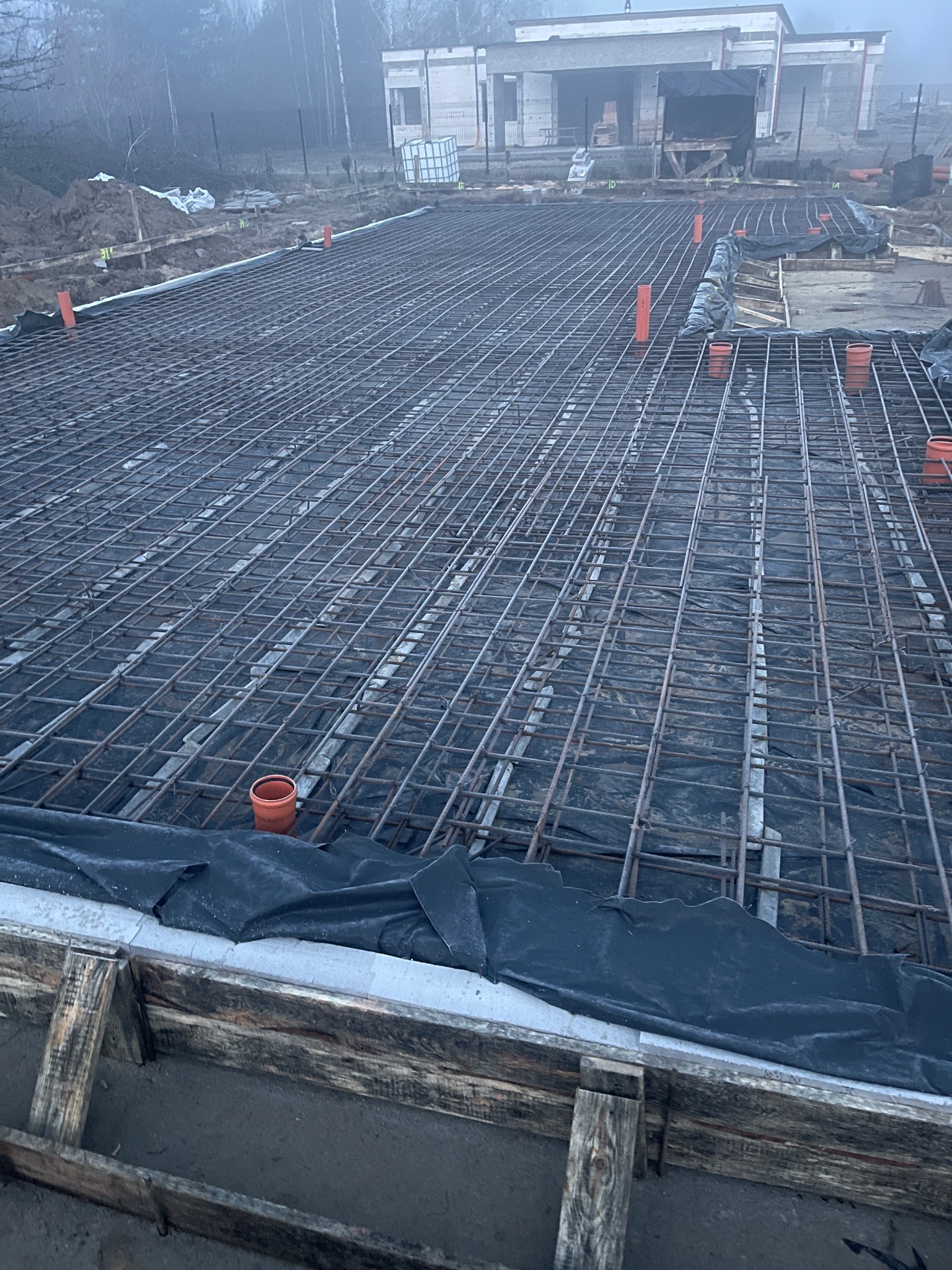 Zbrojenie fundamentu z widoczną siatką stalową, deskowaniem i rurami wentylacyjnymi. W tle budynek w stanie surowym i mglista aura.