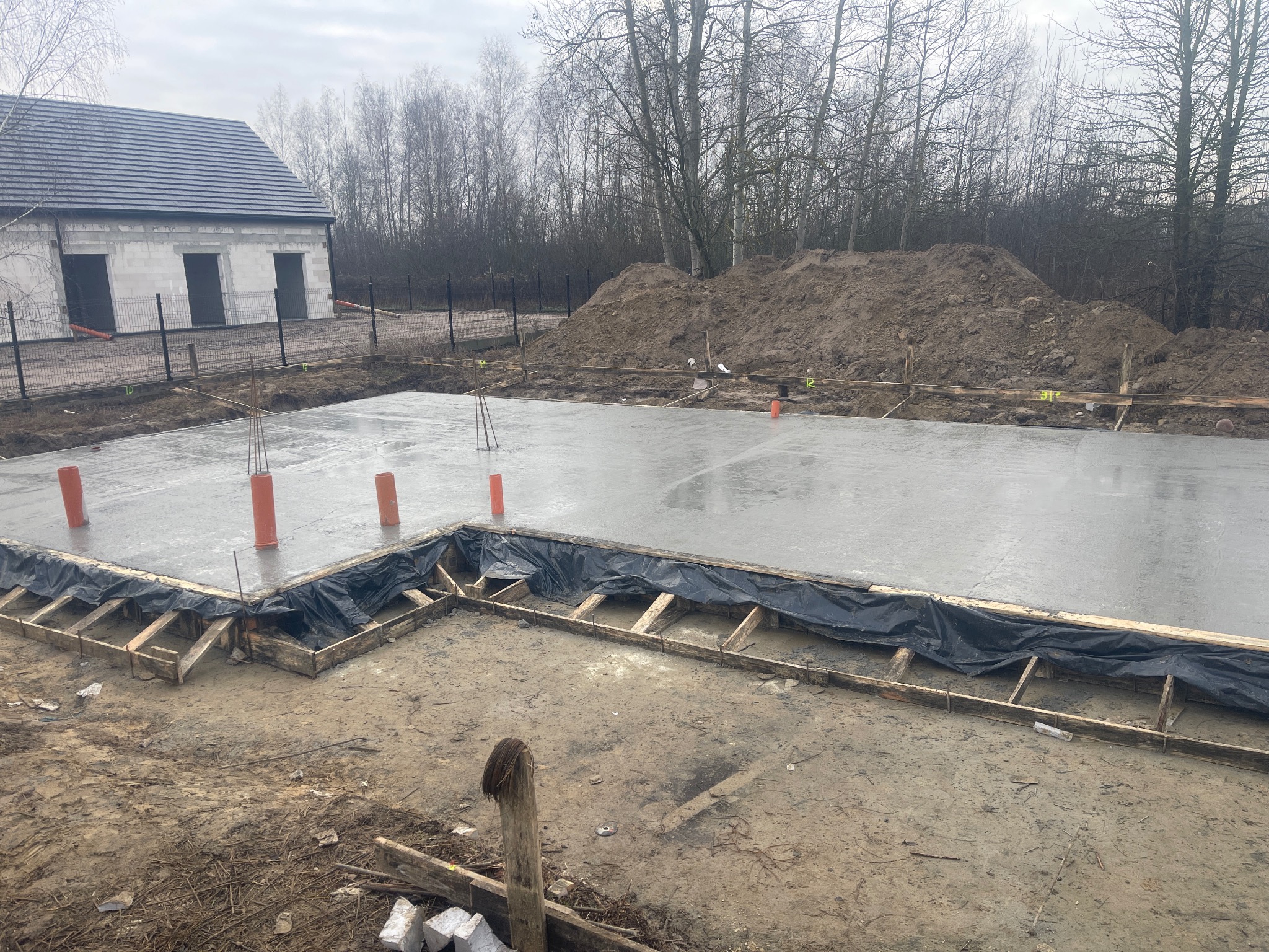 Wylany fundament pod budynek z widocznymi szalunkami i wystającymi rurami wentylacyjnymi. W tle budynek w stanie surowym otwartym i hałda ziemi.