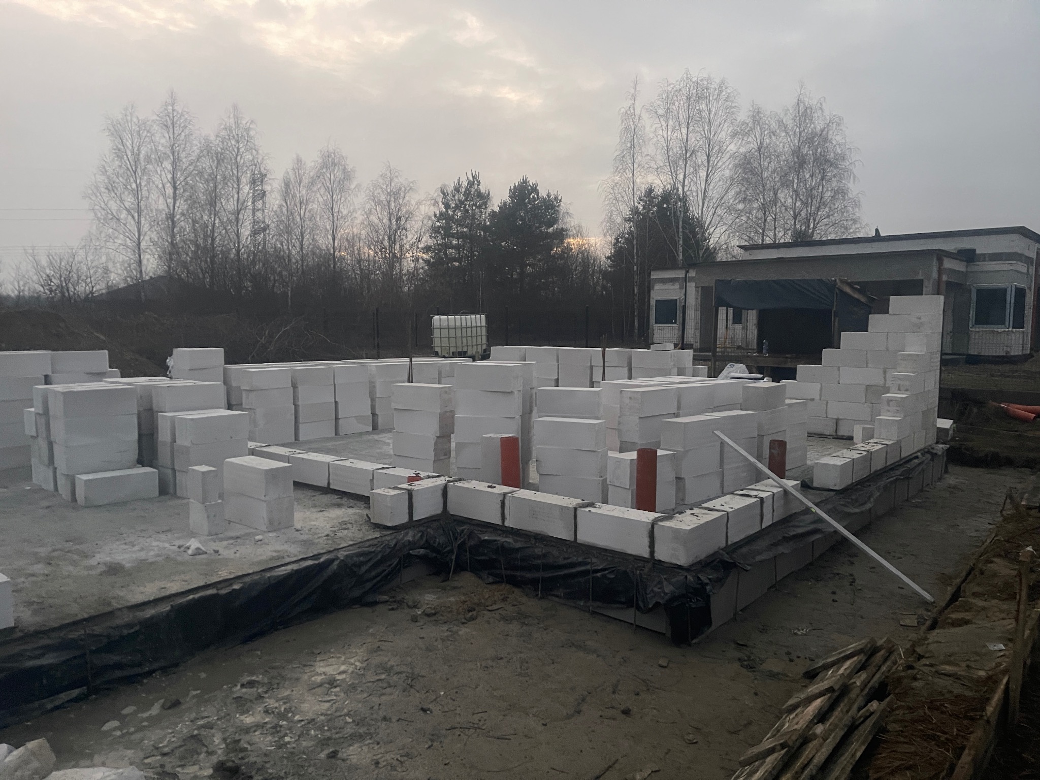 Wznoszenie ścian fundamentowych z białych bloczków na czarnej folii izolacyjnej. Widoczne ułożone bloczki i przygotowane materiały budowlane na placu budowy.