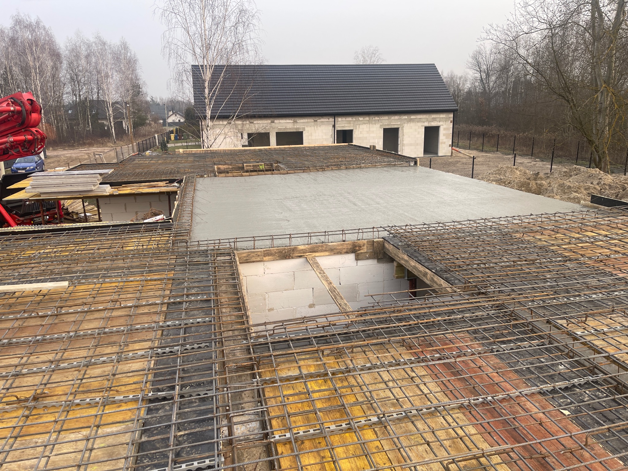 Zbrojenie stropu budynku w trakcie wylewania betonu, widok z góry. W tle surowy budynek z białego pustaka i ciemnym dachem. Na pierwszym planie siatka zbrojeniowa.