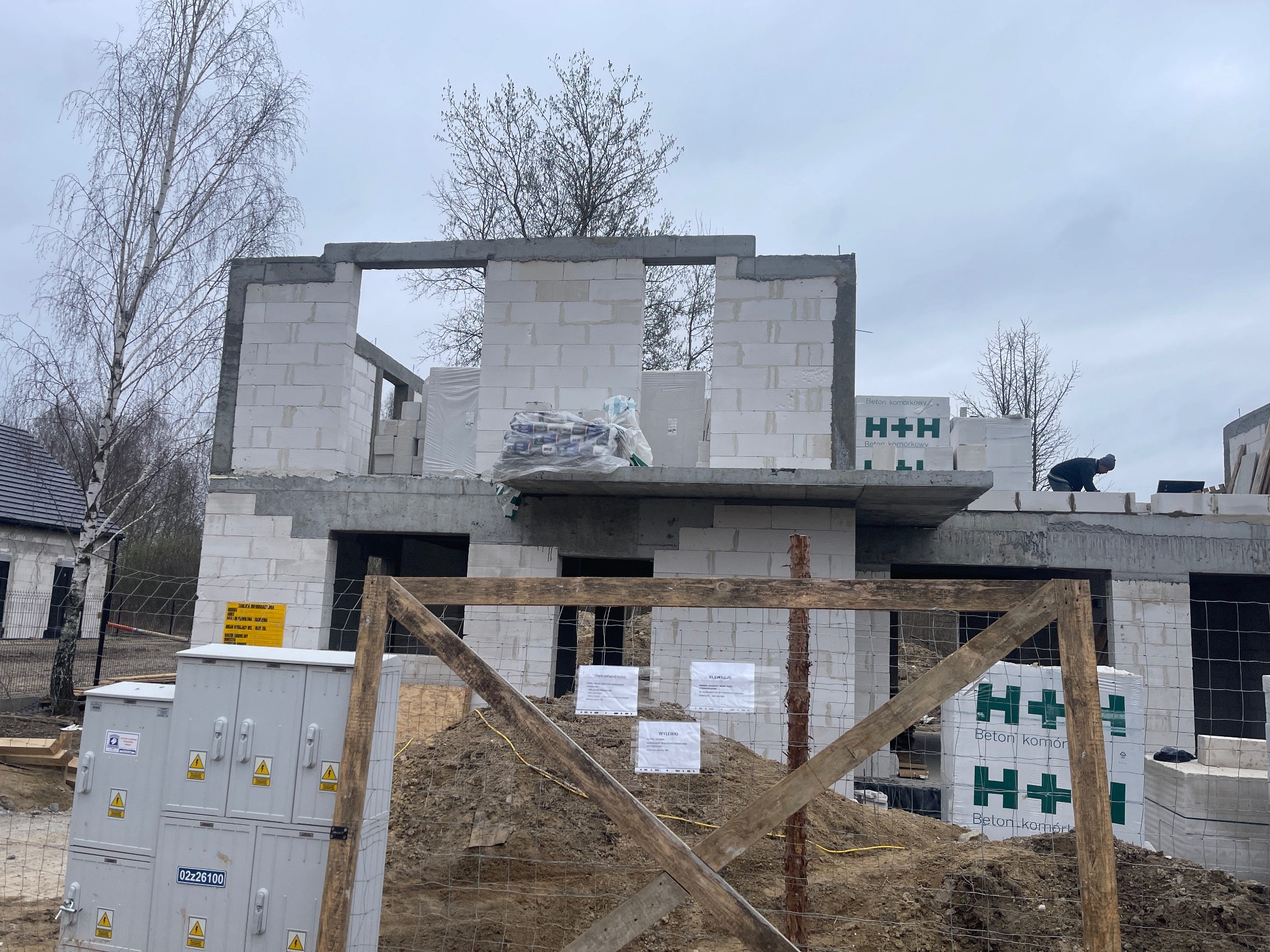 Budowa domu z betonu komórkowego H+H w trakcie realizacji. Widoczne ściany z pustaków, strop, materiały budowlane i tablica informacyjna na ogrodzeniu placu budowy.