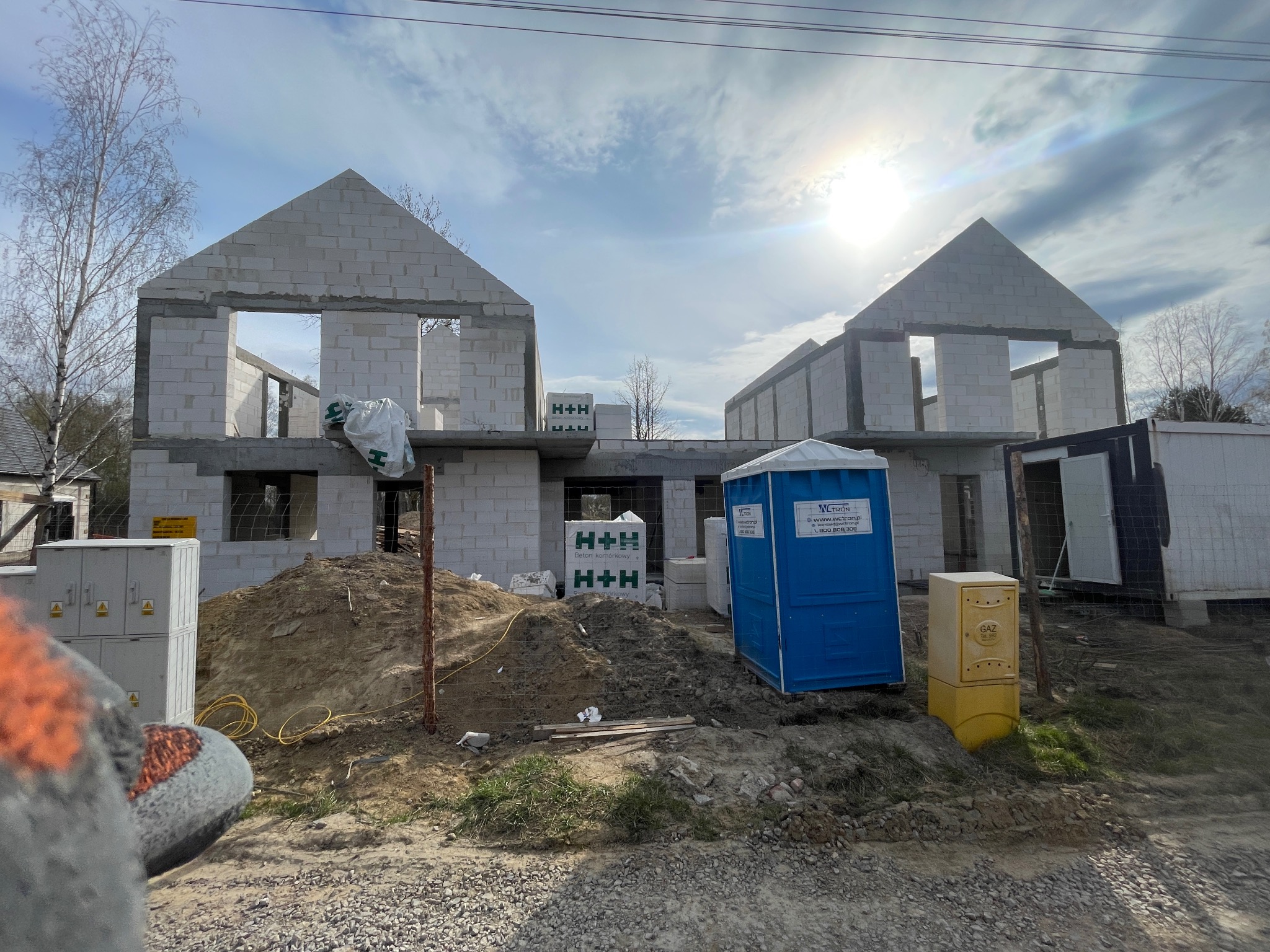 Surowy stan dwóch domów z betonu komórkowego H+H w trakcie budowy, z widocznymi otworami okiennymi i dachami w kształcie litery A, na tle błękitnego nieba.