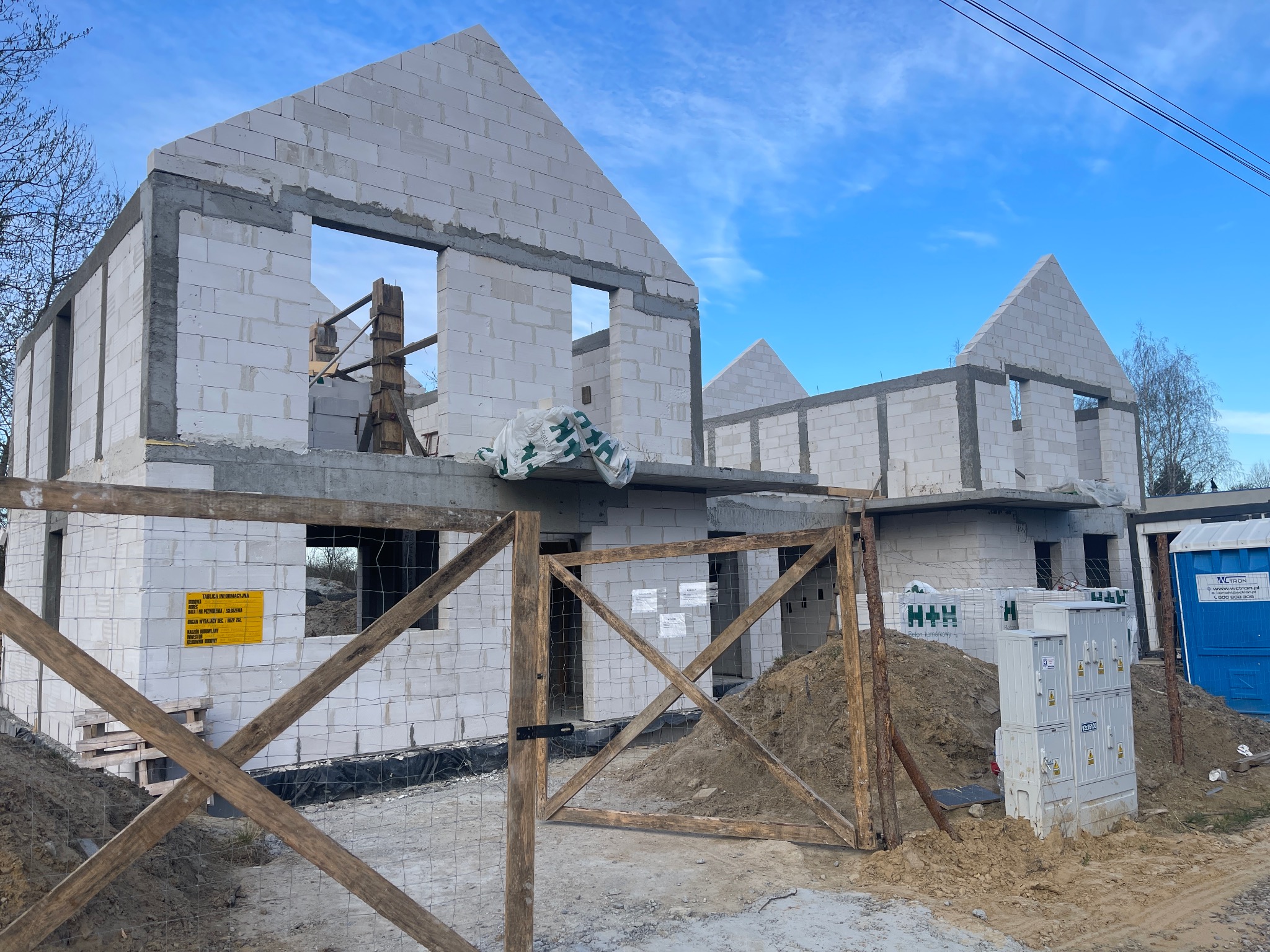 Dwa domy w stanie surowym otwartym, murowane z białych bloczków, widoczne przez drewniane ogrodzenie z siatką. Na pierwszym planie skrzynka elektryczna i toaleta przenośna.