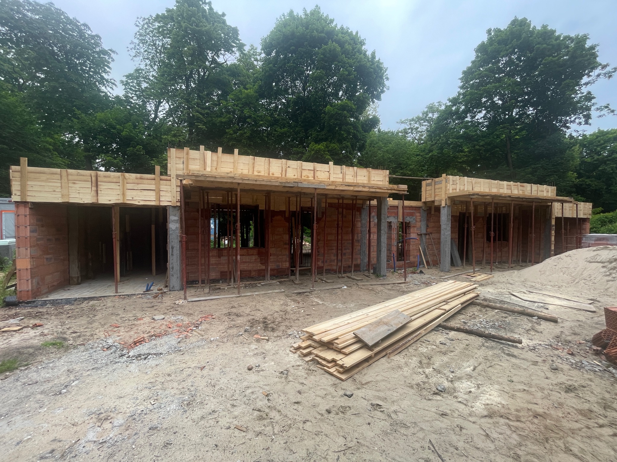 Wznoszenie parterowego domu z czerwonej cegły. Widoczne drewniane szalunki stropowe i stos desek na piaszczystym podłożu. W tle zieleń drzew.