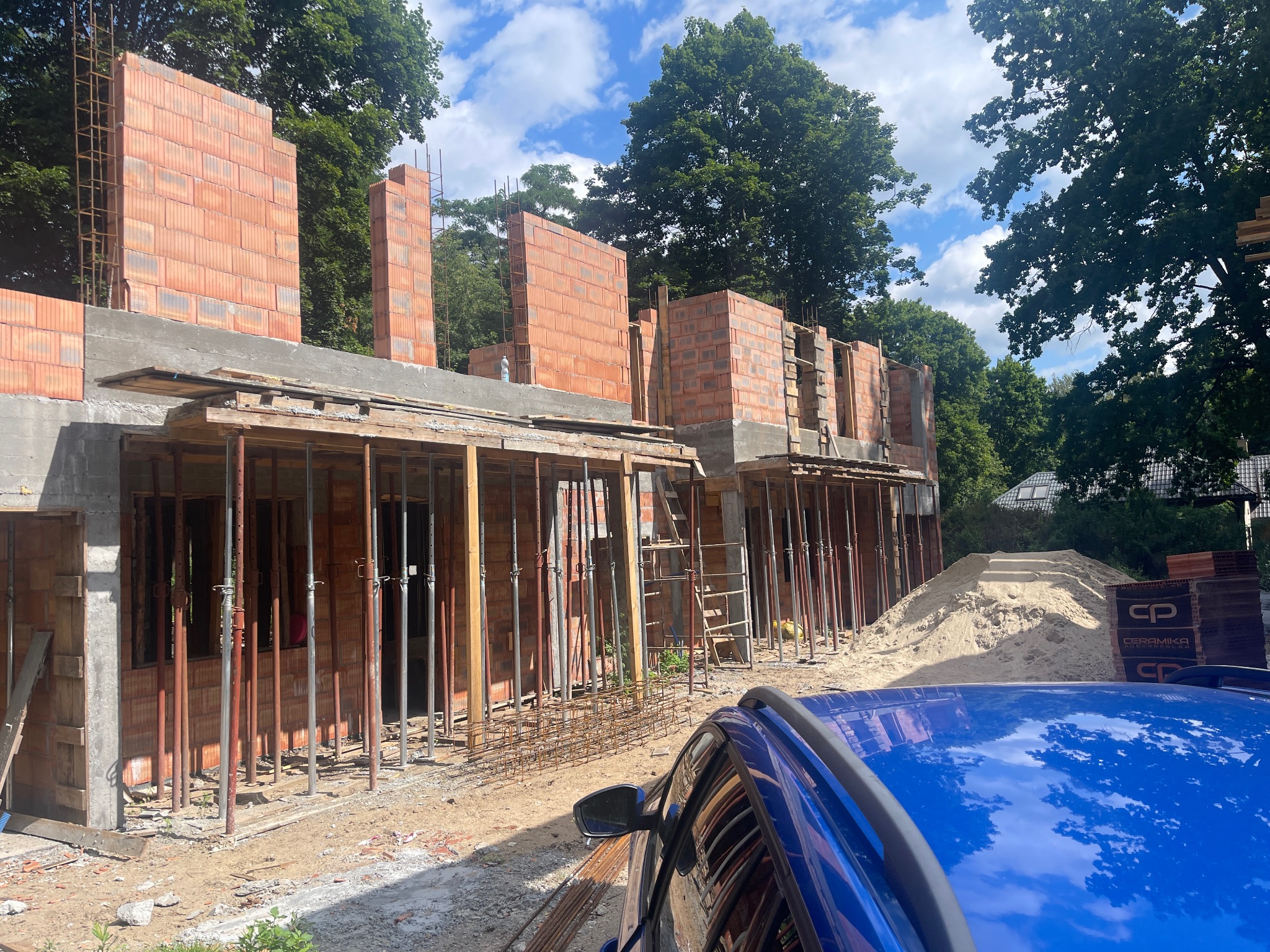 Wznoszenie ścian z czerwonej cegły na budowie domu jednorodzinnego. Widoczne podpory szalunkowe i zbrojenia. W tle drzewa i błękitne niebo z chmurami.