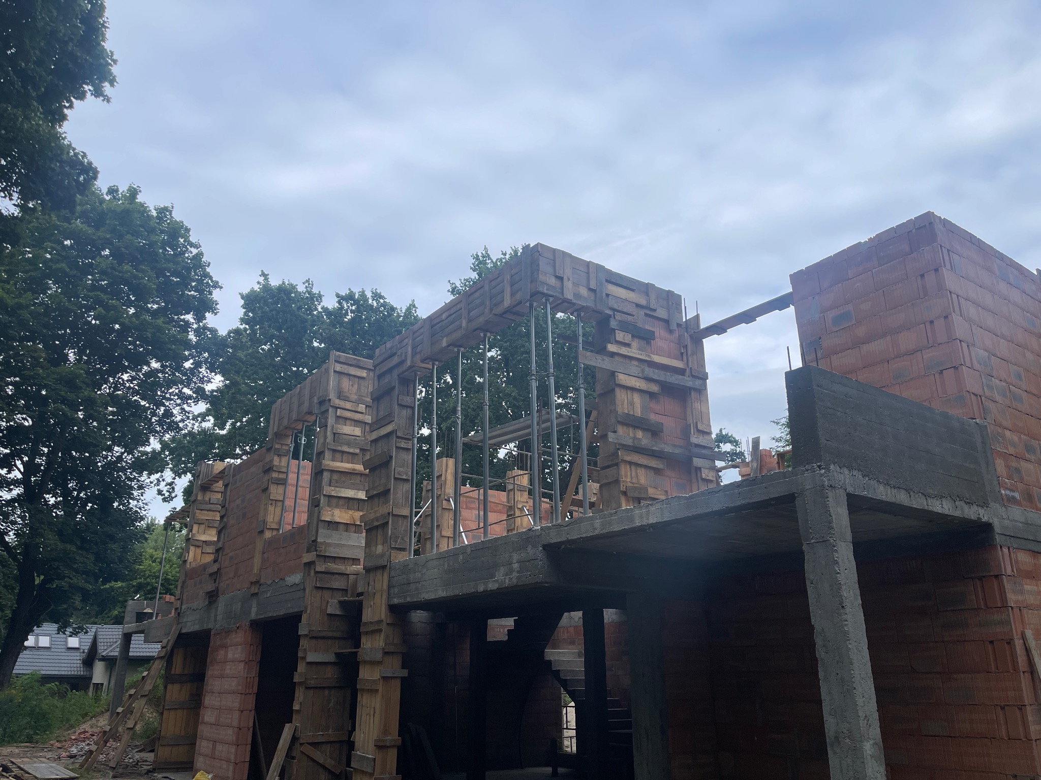Wznoszenie ścian domu z pustaków ceramicznych, z widocznym szalunkiem drewnianym i betonowymi elementami konstrukcyjnymi. W tle zieleń drzew i pochmurne niebo.