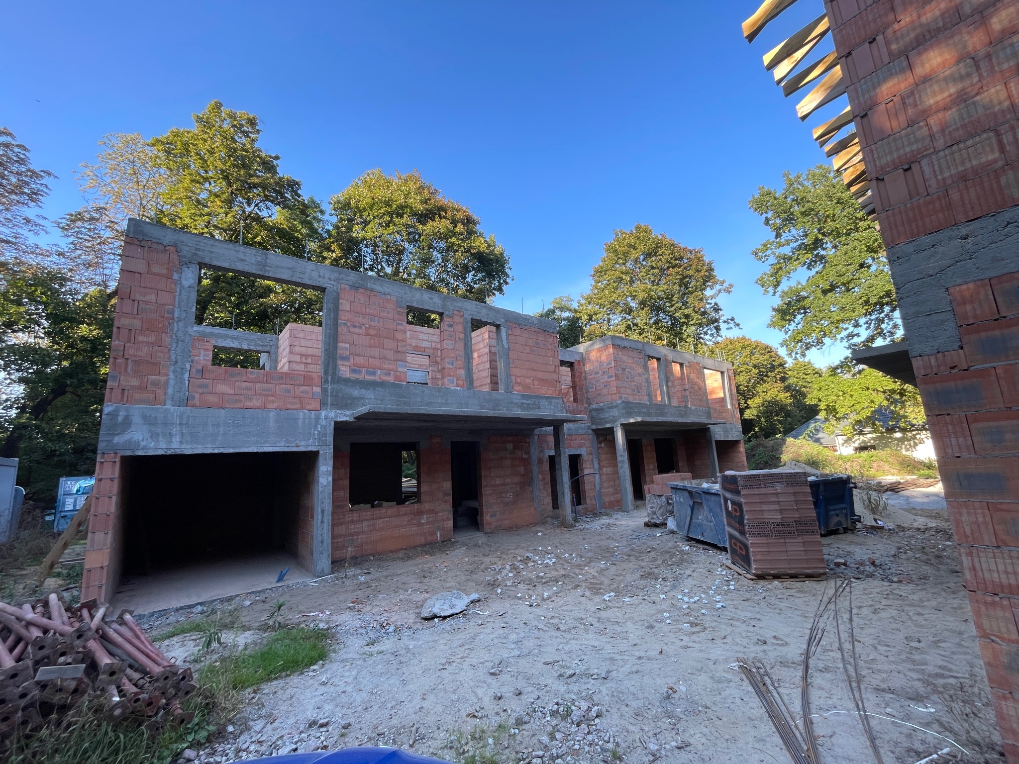 Surowy stan dwóch budynków z czerwonej cegły, z widocznymi otworami okiennymi i betonowymi nadprożami, na tle zielonych drzew i błękitnego nieba.