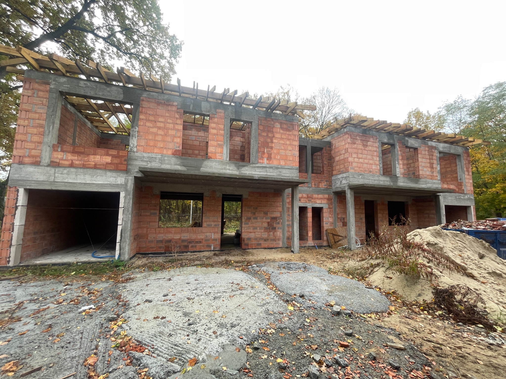 Surowy stan dwukondygnacyjnego domu z czerwonej cegły, z widocznymi drewnianymi elementami konstrukcji dachu i betonowymi nadprożami, na tle jesiennego krajobrazu.