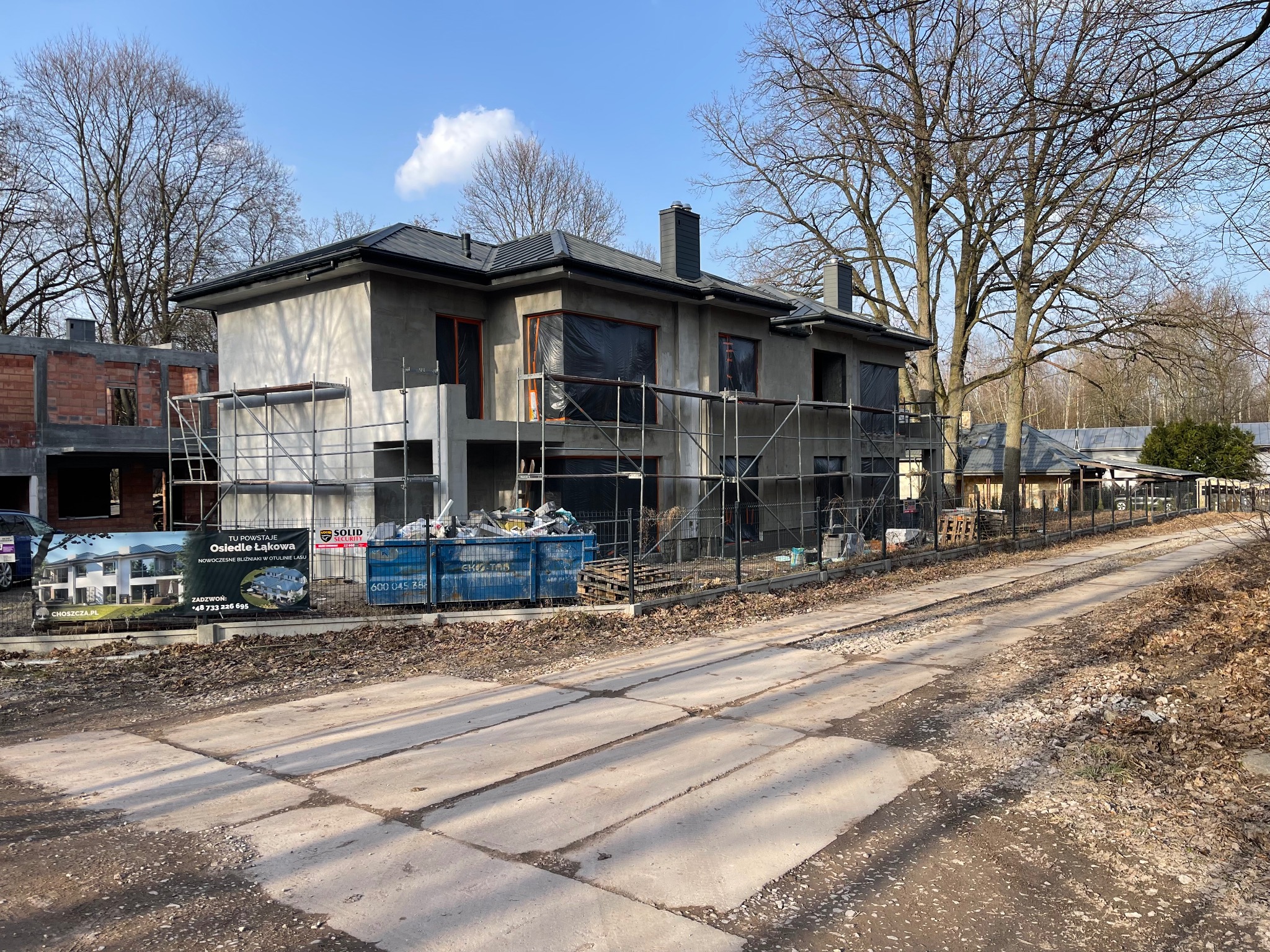 Surowy, dwukondygnacyjny dom w trakcie budowy, z szarym tynkiem i rusztowaniami. W tle drzewa bez liści i baner reklamowy osiedla.