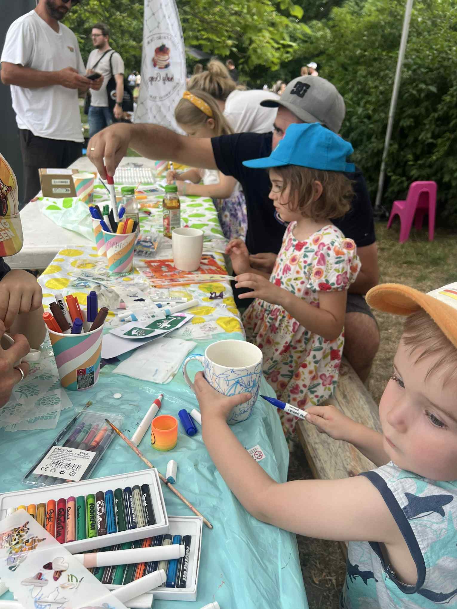 Warsztaty plastyczne dla dzieci: grupa dzieci rysuje markerami na kubkach przy stole z kolorowym obrusem. Widoczne farby, kredki i inne akcesoria.