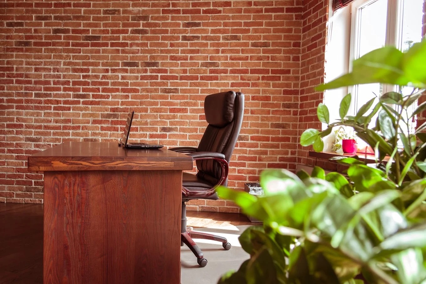 Biuro z ceglaną ścianą, drewnianym biurkiem i laptopem. Krzesło biurowe z wysokim oparciem. Roślina doniczkowa w prawym dolnym rogu. Naturalne światło z okna.