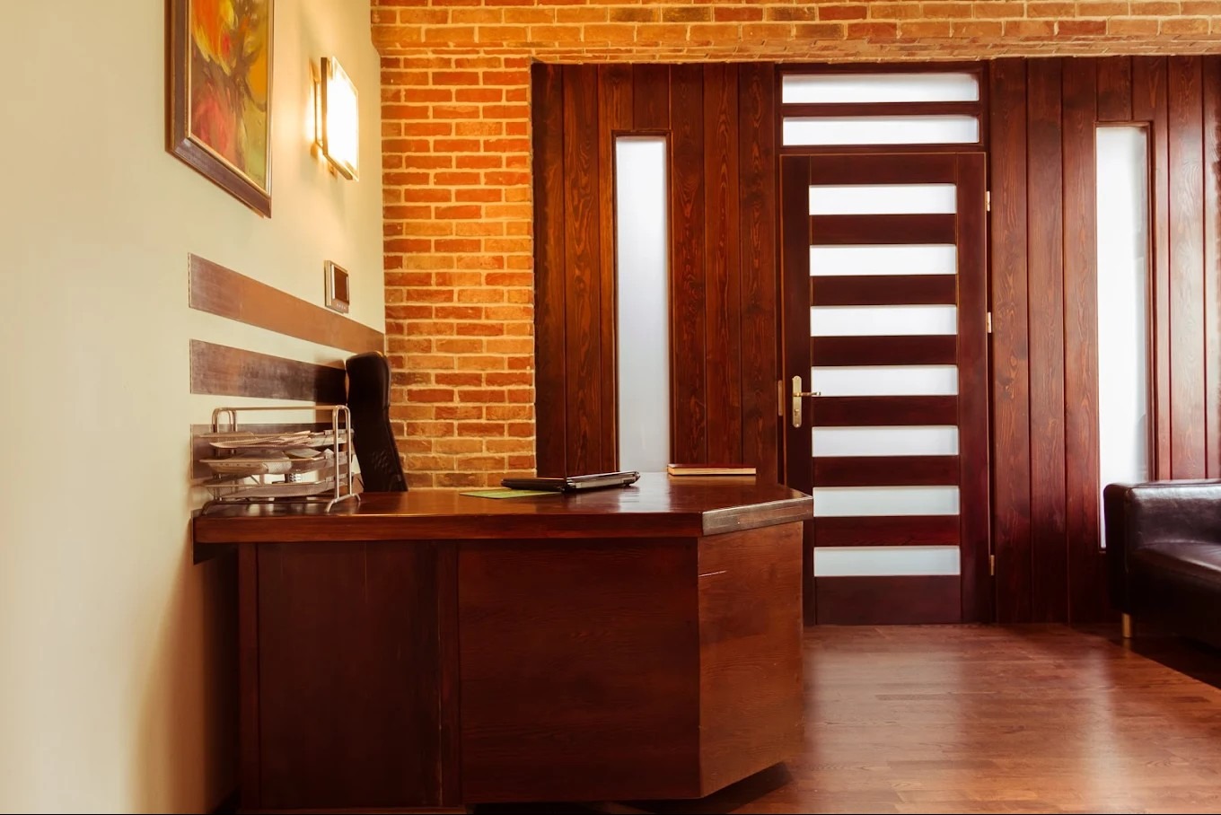 Eleganckie biuro z drewnianym biurkiem, ceglaną ścianą i nowoczesnymi drzwiami z przeszkleniami. Na biurku leży laptop i dokumenty. Ciepłe oświetlenie tworzy przytulną atmosferę.