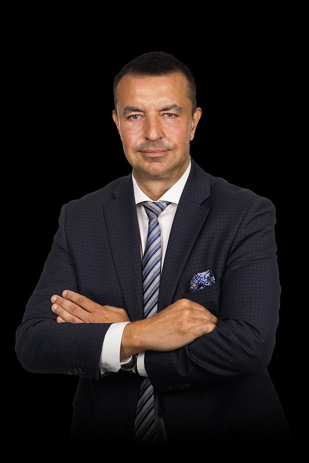 Portret mężczyzny w garniturze na czarnym tle, z założonymi rękami, prezentujący profesjonalny wizerunek.