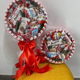 Balonowa Królowa JUSTYNA SŁABOŃSKA - Dwa okrągłe kosze prezentowe z różnymi słodyczami Kinder, owinięte folią i ozdobione czerwonymi kokardami, stoją na żółtym pufie na tle białej ściany.