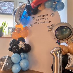 Balonowa Królowa JUSTYNA SŁABOŃSKA - Dekoracja balonowa na roczek w stylu kosmicznym: balony w kształcie rakiety, astronauty, planety i cyfry 1. Na białym tle napis 'Houston we have a one year old'.