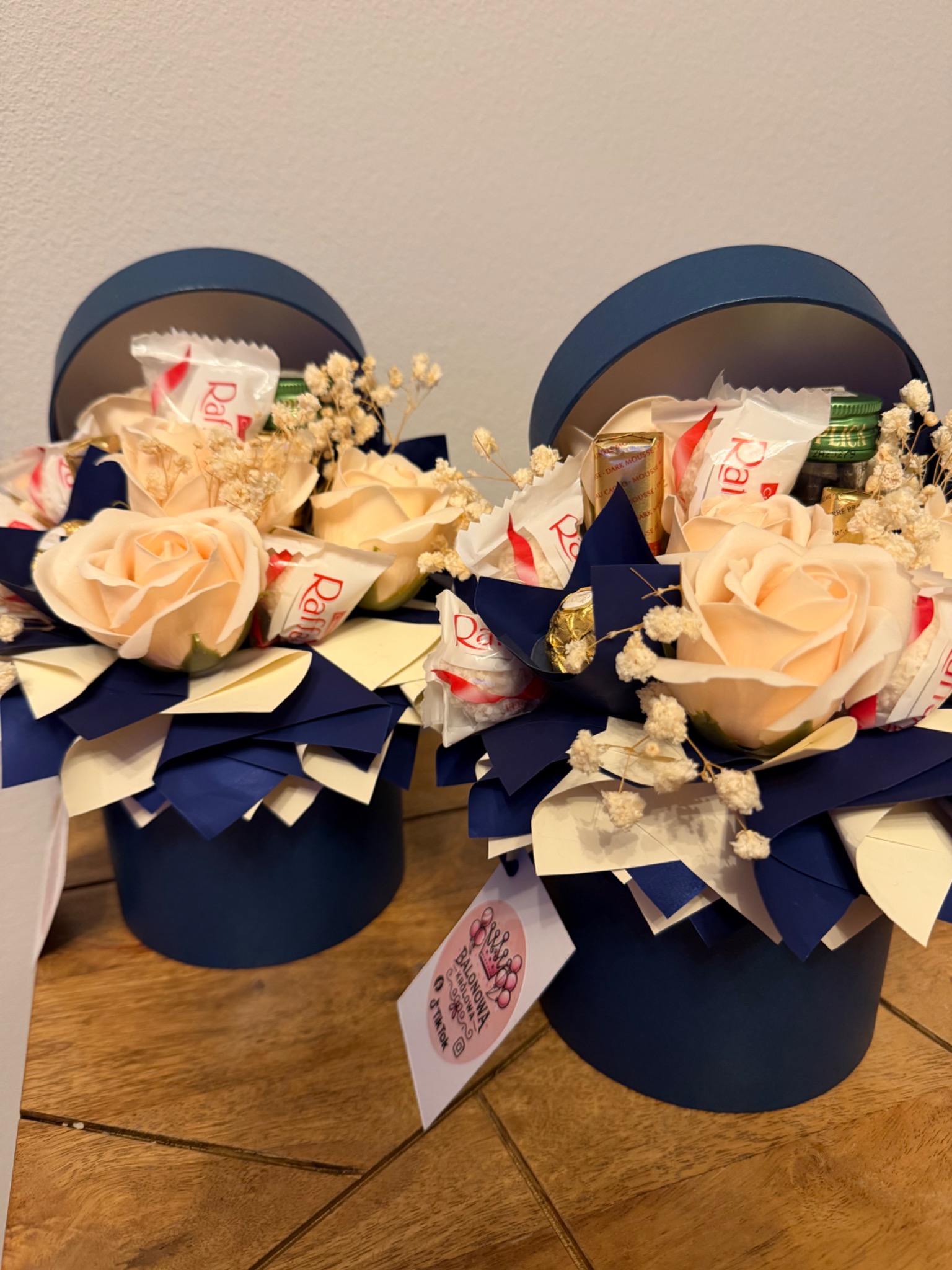 Dwa granatowe pudełka z kwiatami, słodyczami i dekoracjami, tworzące unikalne kompozycje prezentowe na drewnianym blacie. Widoczna etykieta z nazwą firmy.