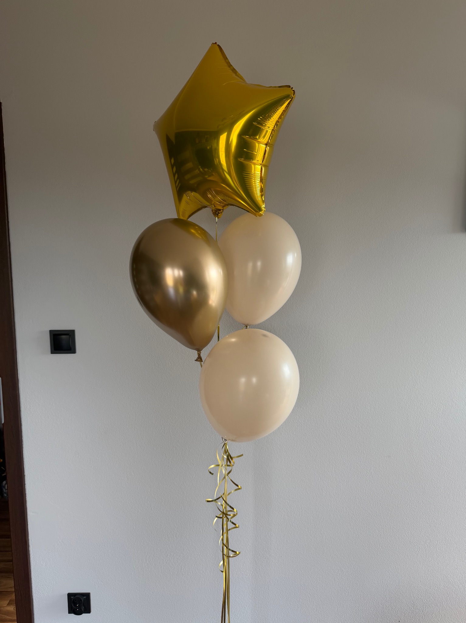 Zestaw balonów: złota gwiazda, złoty okrągły i dwa beżowe, z wstążkami. Dekoracja na imprezę, na tle jasnej ściany z włącznikami.