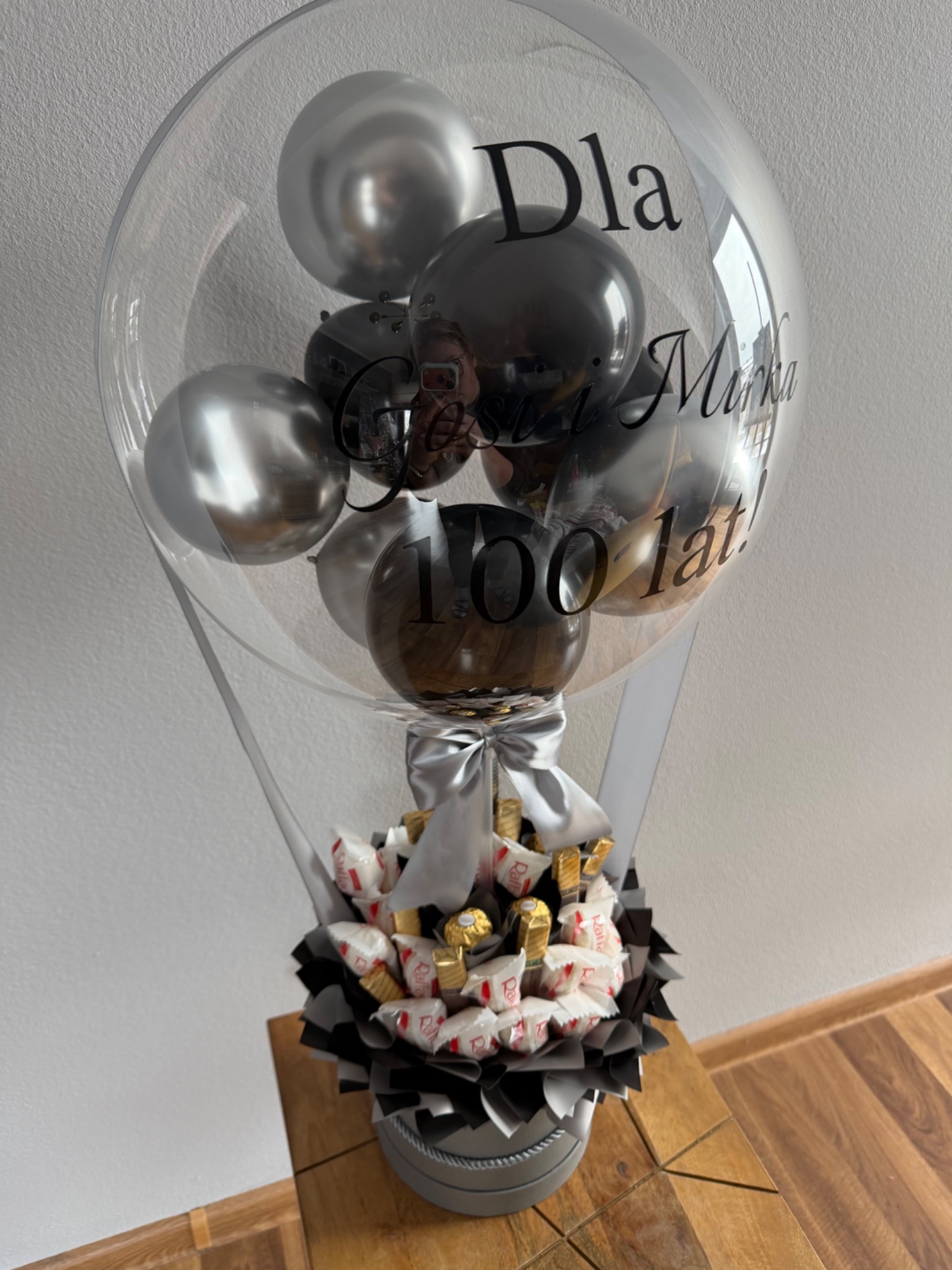 Ozdobny balon z napisem 'Dla Mirka 100 lat!' wypełniony czarno-srebrnymi balonami i bukietem słodyczy, przewiązany srebrną wstążką, na drewnianym stole.