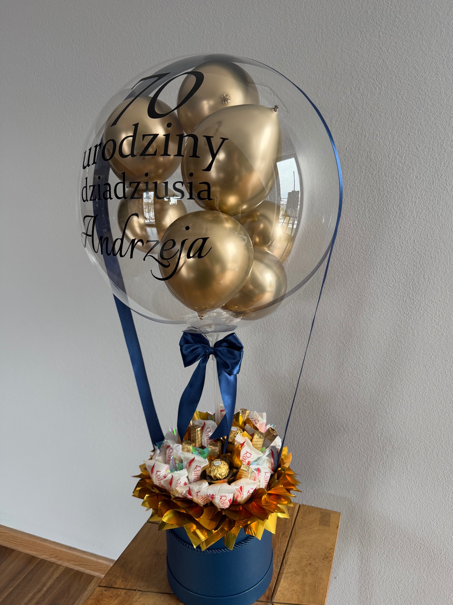 Personalizowany balon z helem i złotymi balonikami w środku, umieszczony w niebieskim pudełku z kompozycją słodyczy, przewiązany niebieską wstążką. Napis na balonie: 70 urodziny dziadziusia Andrzeja.