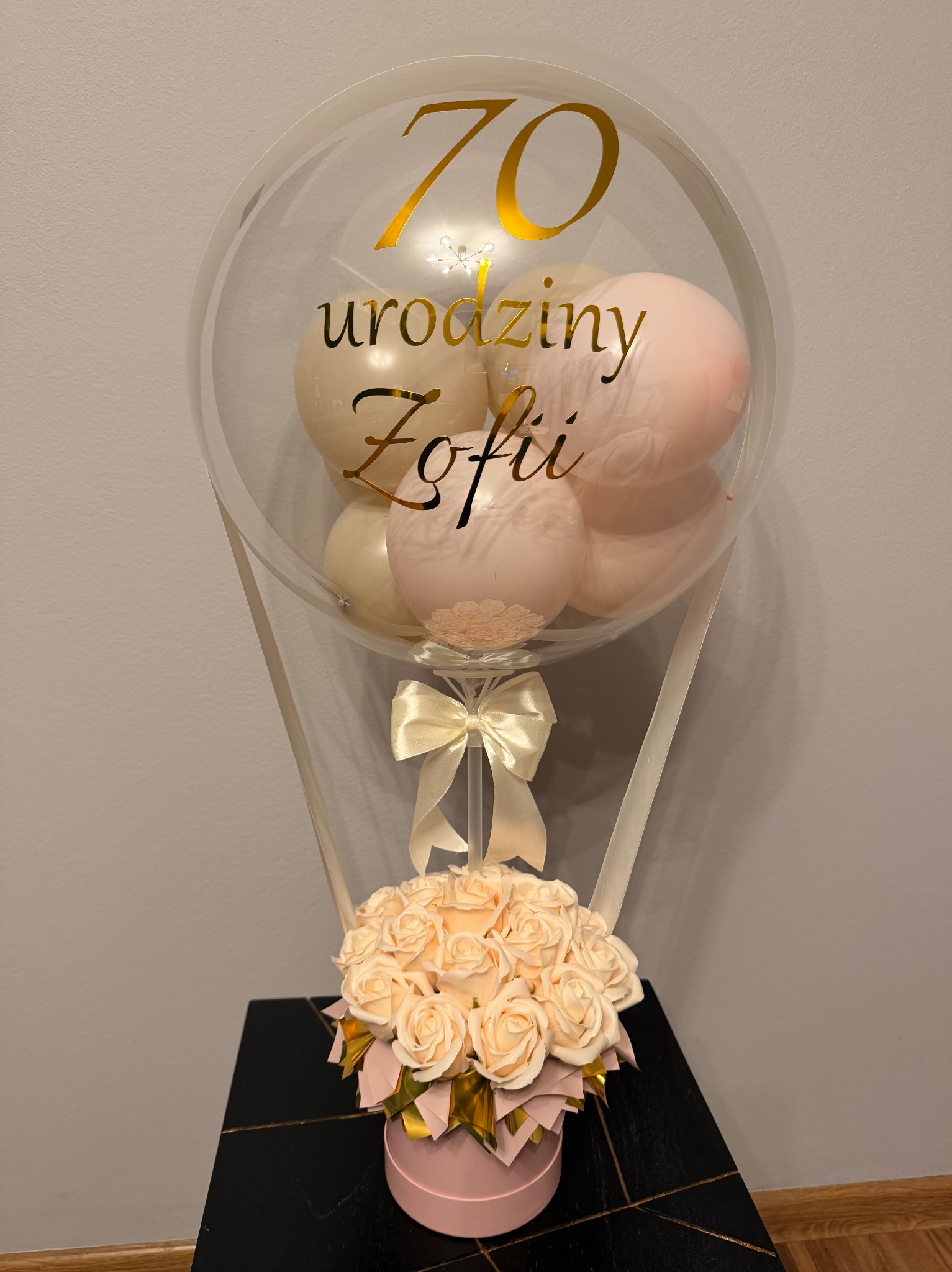 Balon z napisem '70 urodziny Zofii' wypełniony mniejszymi balonami, umieszczony nad bukietem kremowych róż w różowym pudełku, całość na czarnym stoliku.