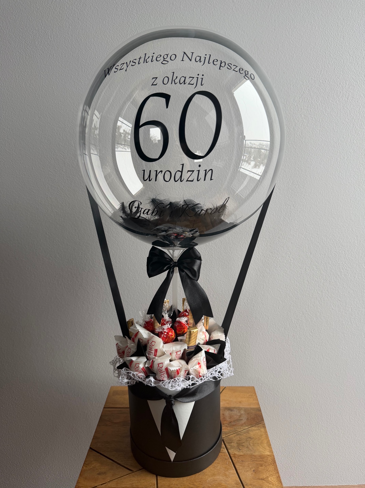 Oryginalny kosz prezentowy na 60 urodziny: balon z napisem, piórka, słodycze Raffaello i Merci w eleganckim pudełku w stylu smokingu, na drewnianym blacie.