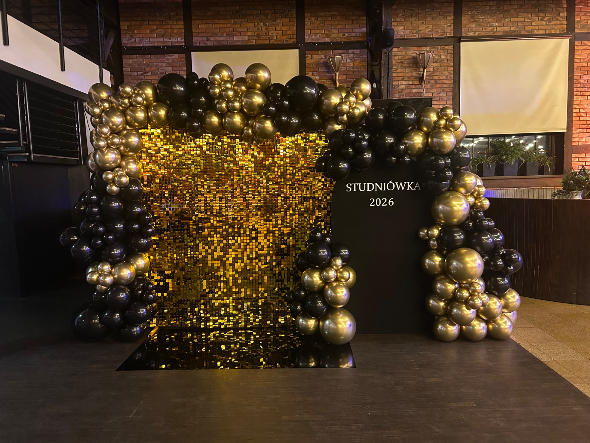Dekoracja studniówkowa 2026: złota ścianka z cekinów otoczona girlandą z czarno-złotych balonów. Elegancki wystrój imprezy w ciemnej sali.