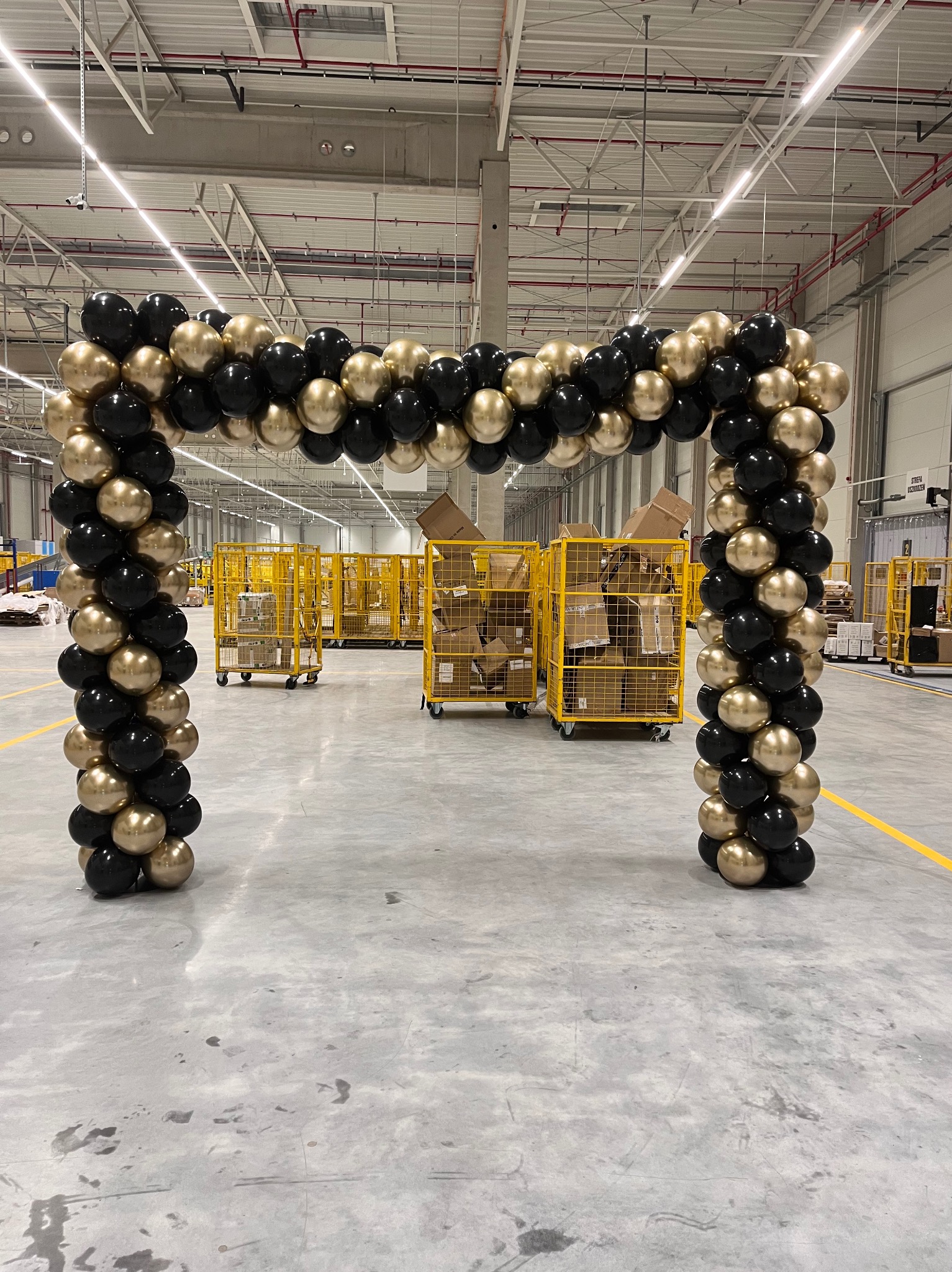 Dekoracja z czarno-złotych balonów w kształcie łuku, ustawiona w hali magazynowej z żółtymi wózkami transportowymi w tle. Dekoracja eventowa.