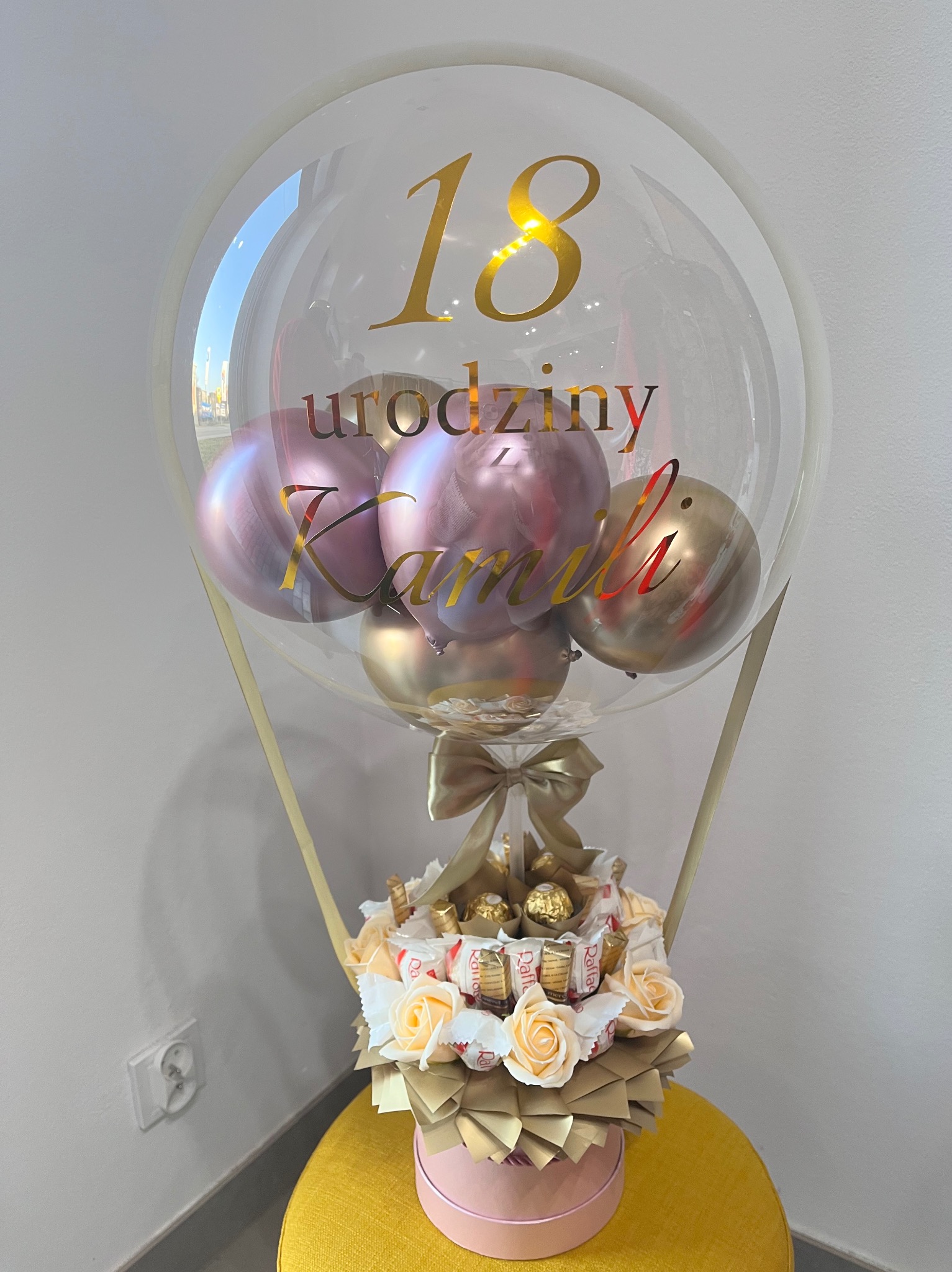 Urodzinowy balon z napisem '18 urodziny Kamili', wypełniony różowymi i złotymi balonami, umieszczony na kompozycji ze słodyczy, róż i złotych elementów dekoracyjnych.