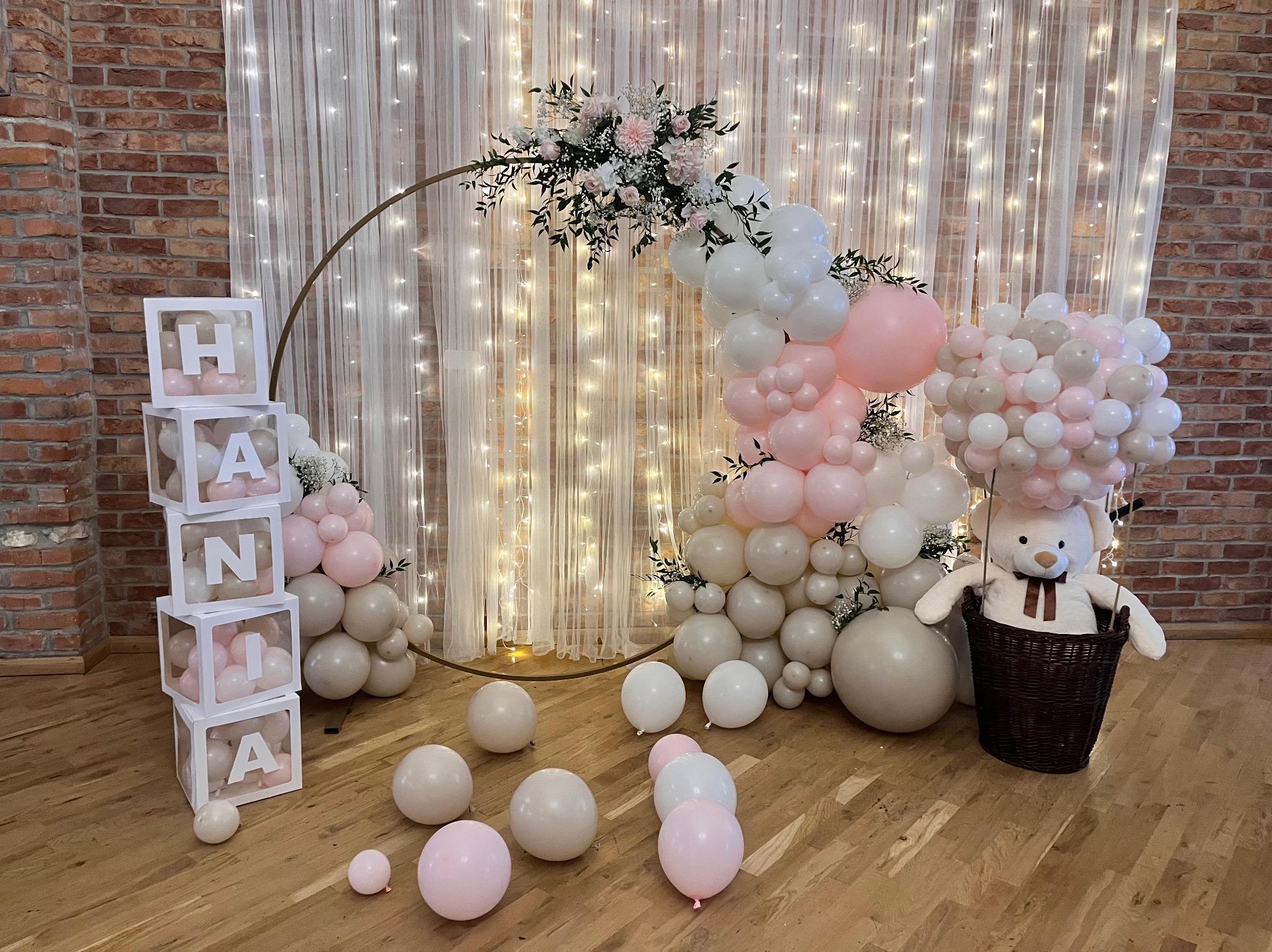 Dekoracja urodzinowa z balonów w odcieniach różu i bieli, z imieniem 'Hania' ułożonym z pudełek, pluszowy miś w koszu, na tle ceglanej ściany i kurtyny świetlnej.