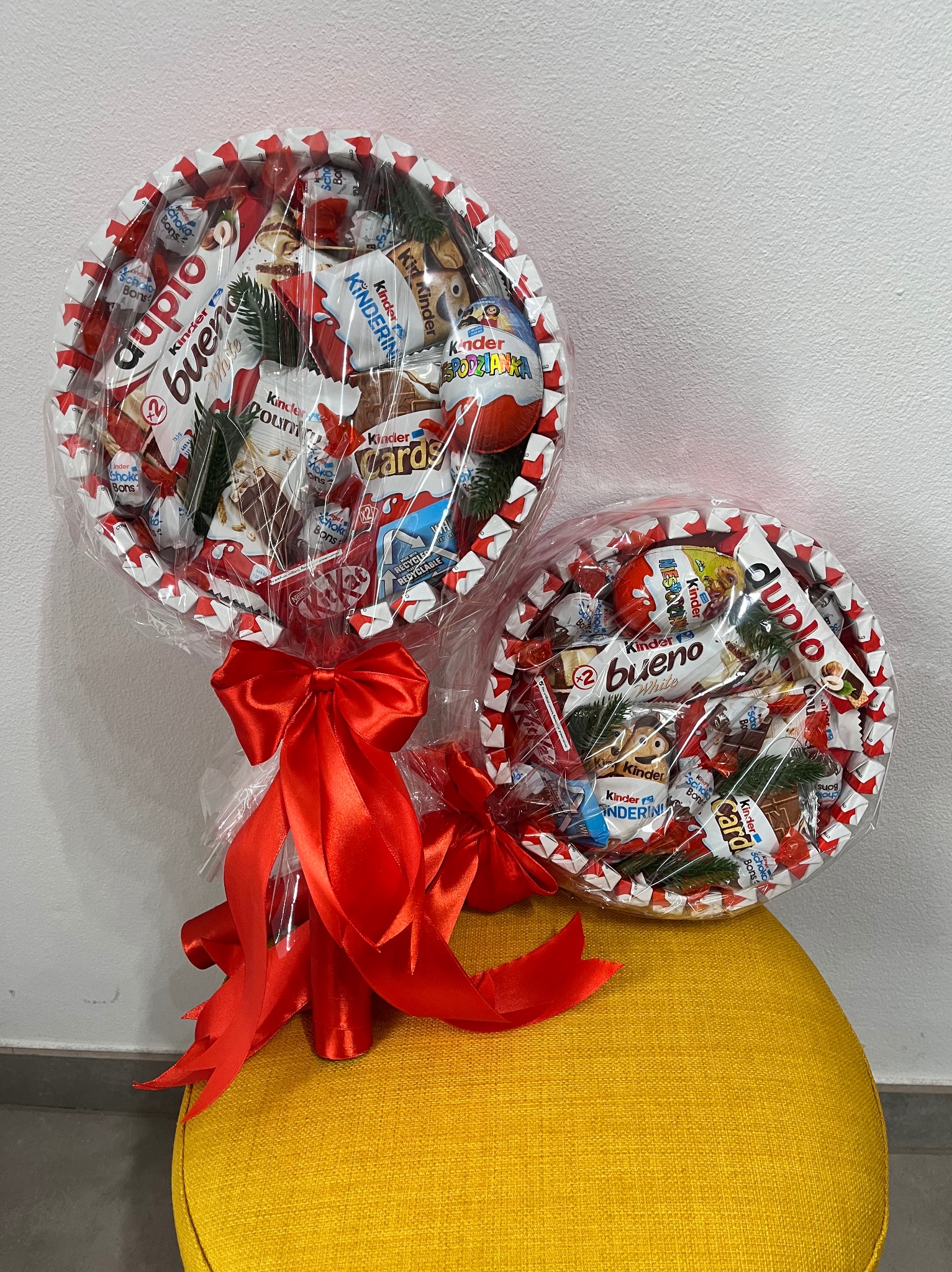 Dwa okrągłe kosze prezentowe z różnymi słodyczami Kinder, owinięte folią i ozdobione czerwonymi kokardami, stoją na żółtym pufie na tle białej ściany.