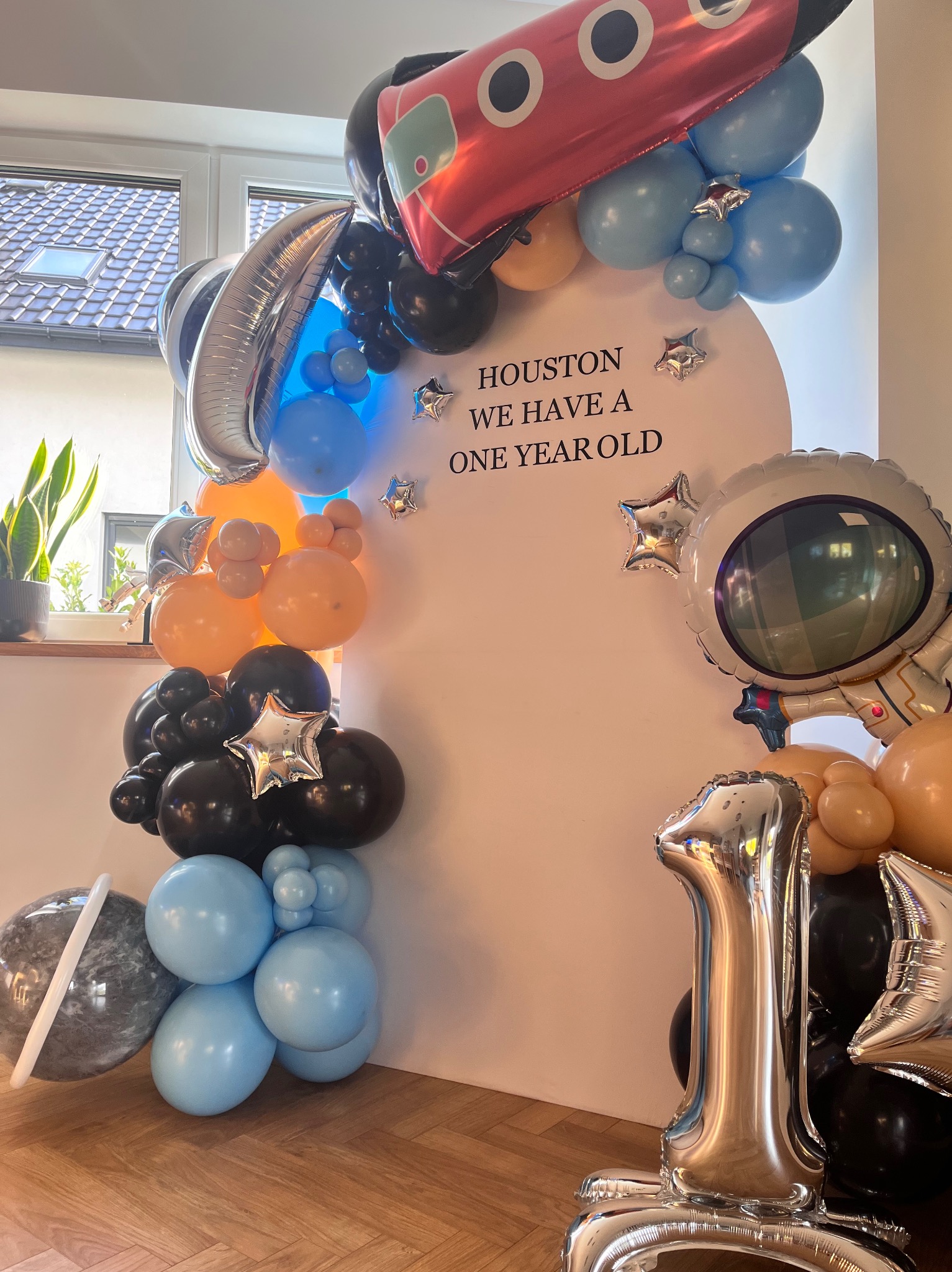 Dekoracja balonowa na roczek w stylu kosmicznym: balony w kształcie rakiety, astronauty, planety i cyfry 1. Na białym tle napis 'Houston we have a one year old'.