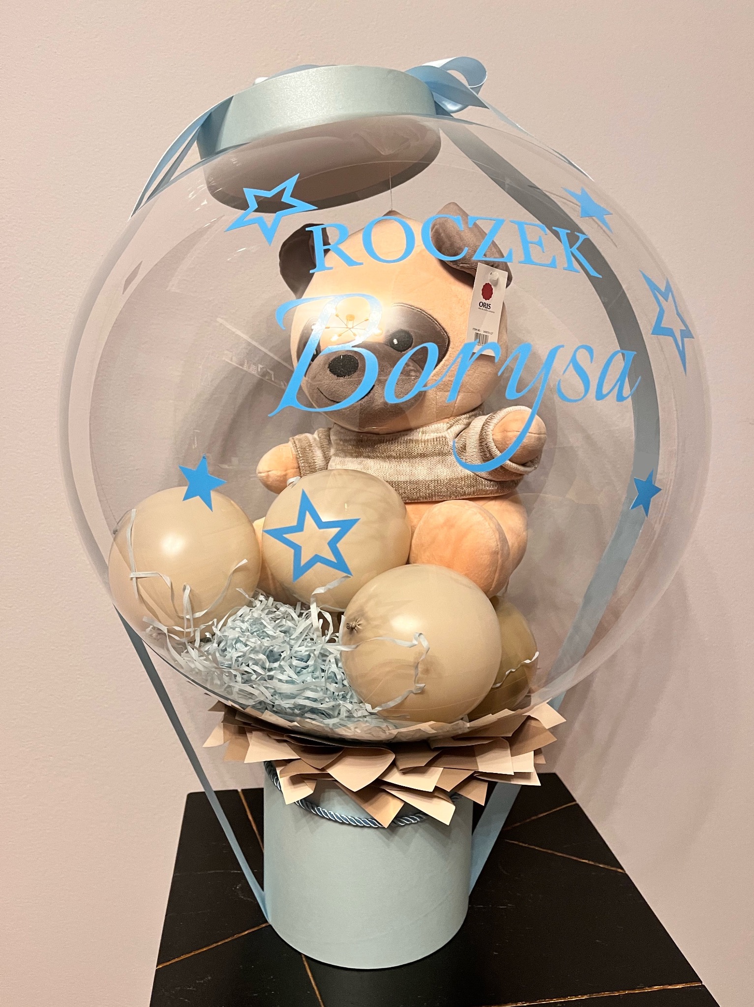 Uroczy prezent na roczek: pluszowy miś w przezroczystym balonie z napisem 'Roczek Borysa', otoczony pastelowymi balonami i dekoracyjnym wypełnieniem.