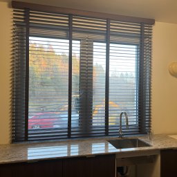 Projekt Meble - Ciemne żaluzje drewniane w oknie kuchennym, widok na zewnątrz z budową. Blat granitowy odbija światło, nowoczesna armatura. Oświetlenie punktowe na ścianie.