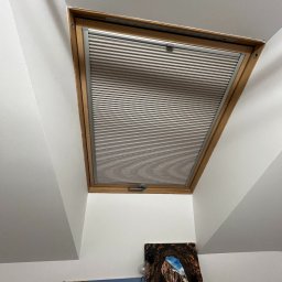 Projekt Meble - Zamontowana plisa okienna w drewnianej ramie okna dachowego, widok z dołu, z dekoracjami ściennymi w tle. Plisa w kolorze szarym, poziome paski.