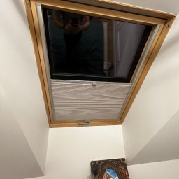 Projekt Meble - Okno dachowe z drewnianą ramą i zamontowaną plisą w kolorze jasnego drewna, widok od dołu, z odbiciem osoby robiącej zdjęcie w szybie.