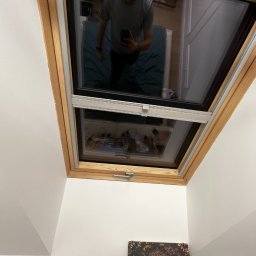 Projekt Meble - Zamknięte okno dachowe z drewnianą ramą i zamontowaną roletą wewnętrzną. Widoczne odbicie osoby robiącej zdjęcie. Na ścianie obrazy.