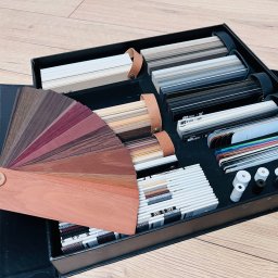 ADRIAN ANTOSZ - Projekt Meble - Paleta kolorów i materiałów do rolet i żaluzji w eleganckim, czarnym pudełku prezentacyjnym, ukazująca różnorodność wykończeń i tekstur na drewnianym tle.