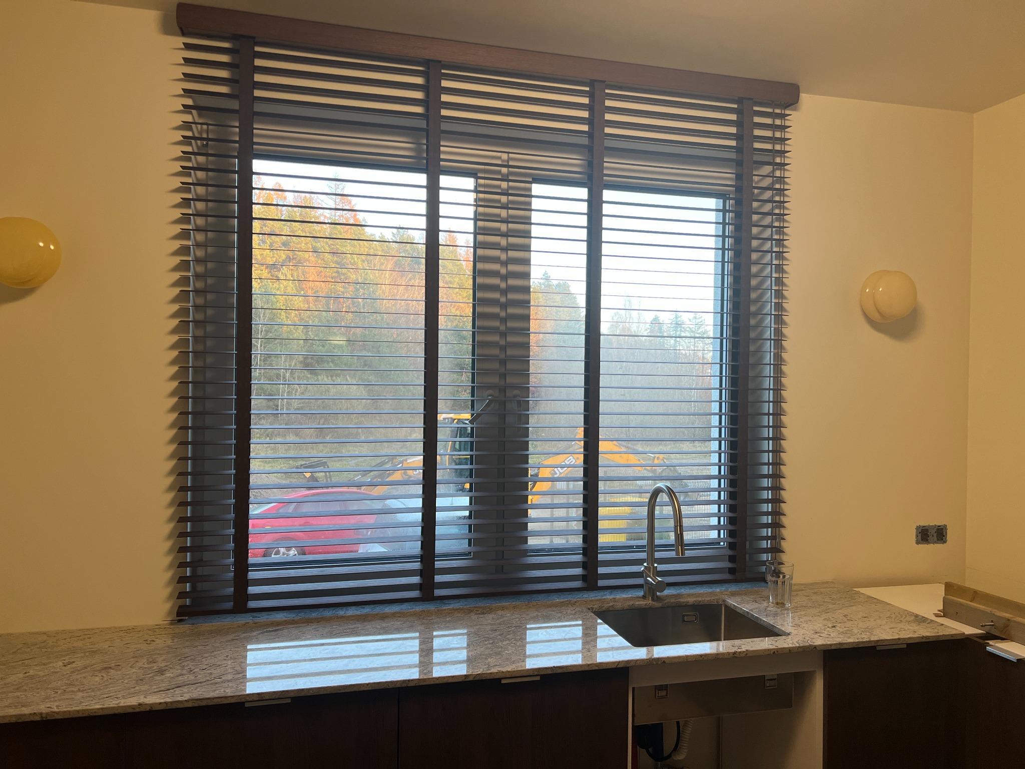 Ciemne żaluzje drewniane w oknie kuchennym, widok na zewnątrz z budową. Blat granitowy odbija światło, nowoczesna armatura. Oświetlenie punktowe na ścianie.