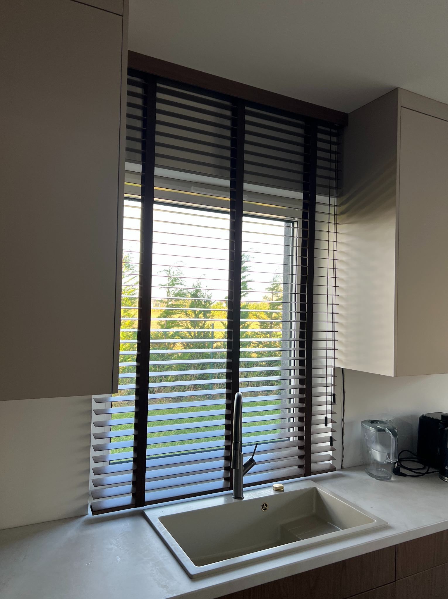 Kuchnia z drewnianymi żaluzjami poziomymi w oknie, widok na zieleń za oknem. Ujęcie z bliska, widoczny zlew i fragment blatu.