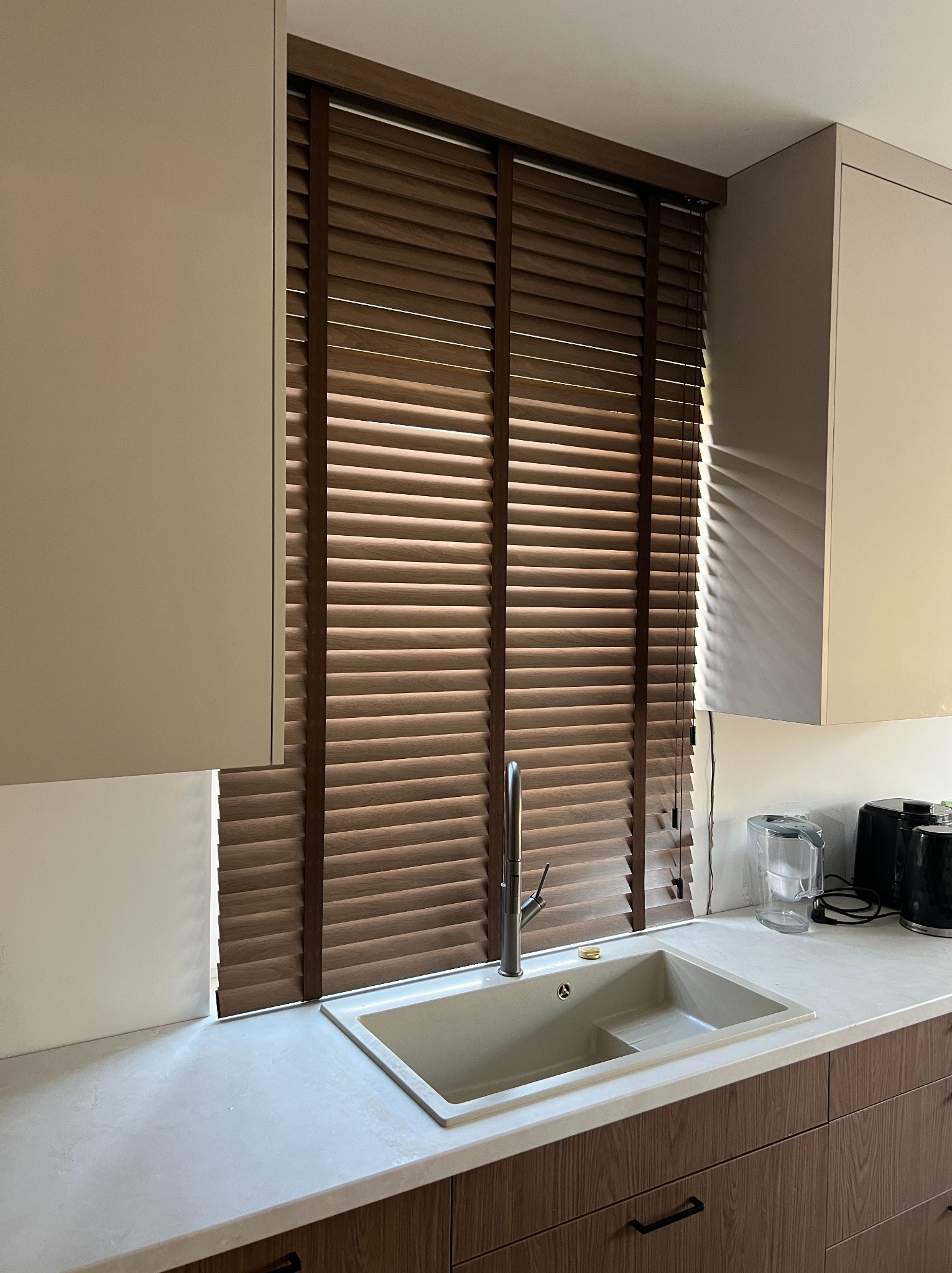 Kuchnia z drewnianymi żaluzjami w oknie, widok na zlew i fragment blatu. Naturalne światło podkreśla strukturę drewna.