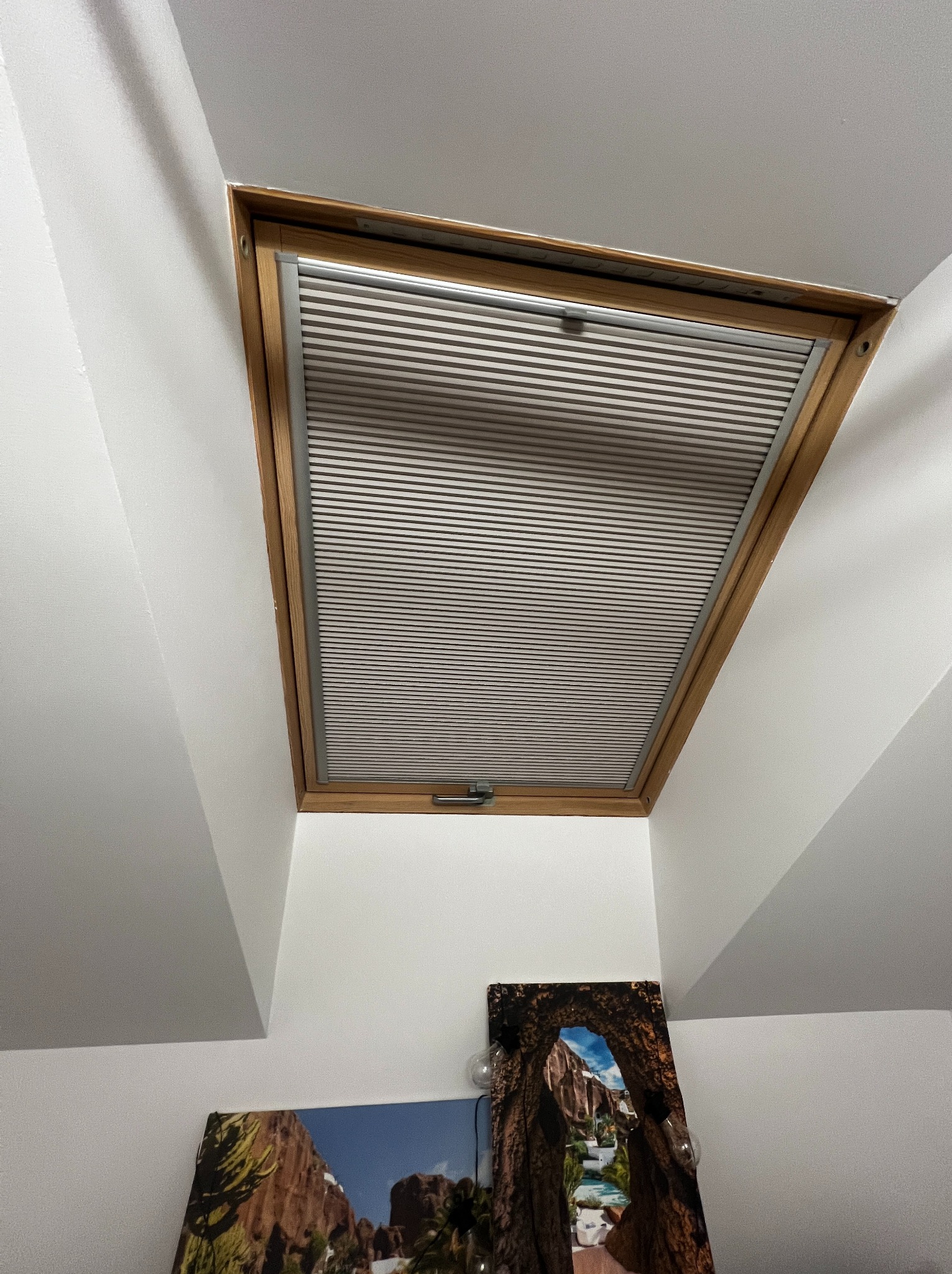 Zamontowana plisa okienna w drewnianej ramie okna dachowego, widok z dołu, z dekoracjami ściennymi w tle. Plisa w kolorze szarym, poziome paski.