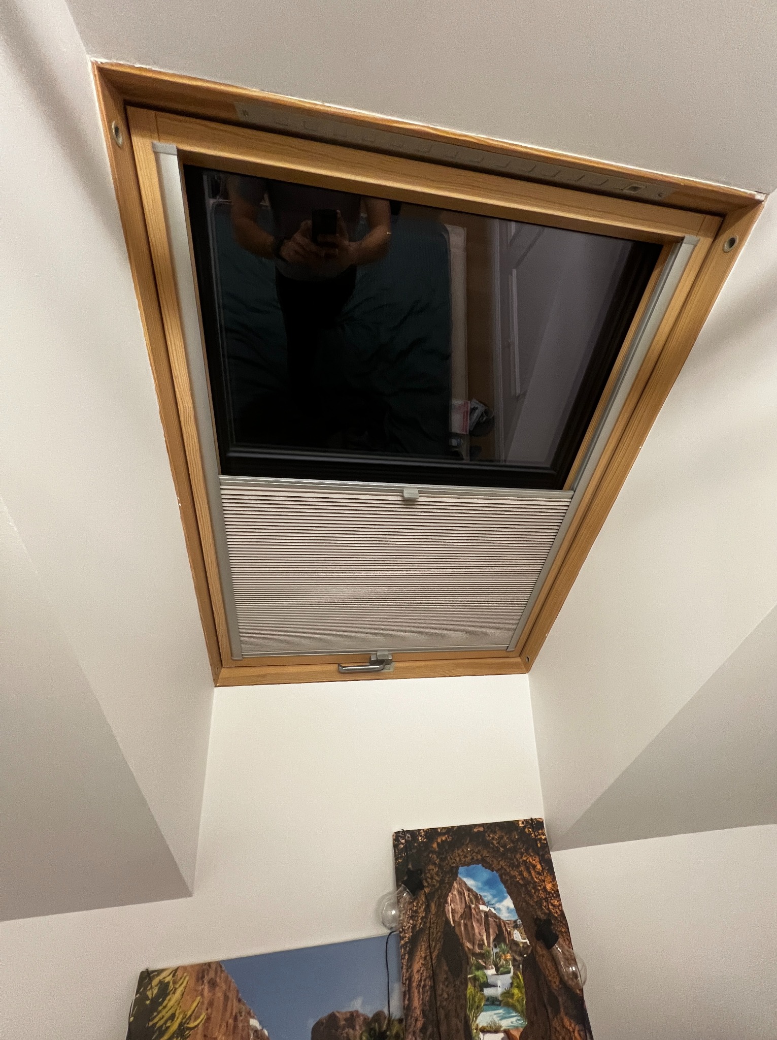 Okno dachowe z drewnianą ramą i zamontowaną plisą w kolorze jasnego drewna, widok od dołu, z odbiciem osoby robiącej zdjęcie w szybie.