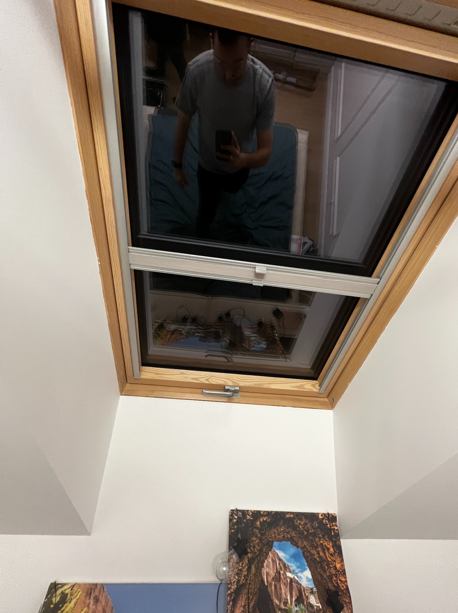 Zamknięte okno dachowe z drewnianą ramą i zamontowaną roletą wewnętrzną. Widoczne odbicie osoby robiącej zdjęcie. Na ścianie obrazy.