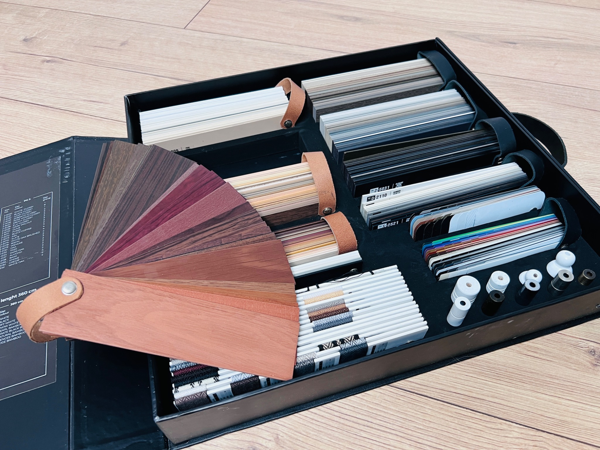 Paleta kolorów i materiałów do rolet i żaluzji w eleganckim, czarnym pudełku prezentacyjnym, ukazująca różnorodność wykończeń i tekstur na drewnianym tle.