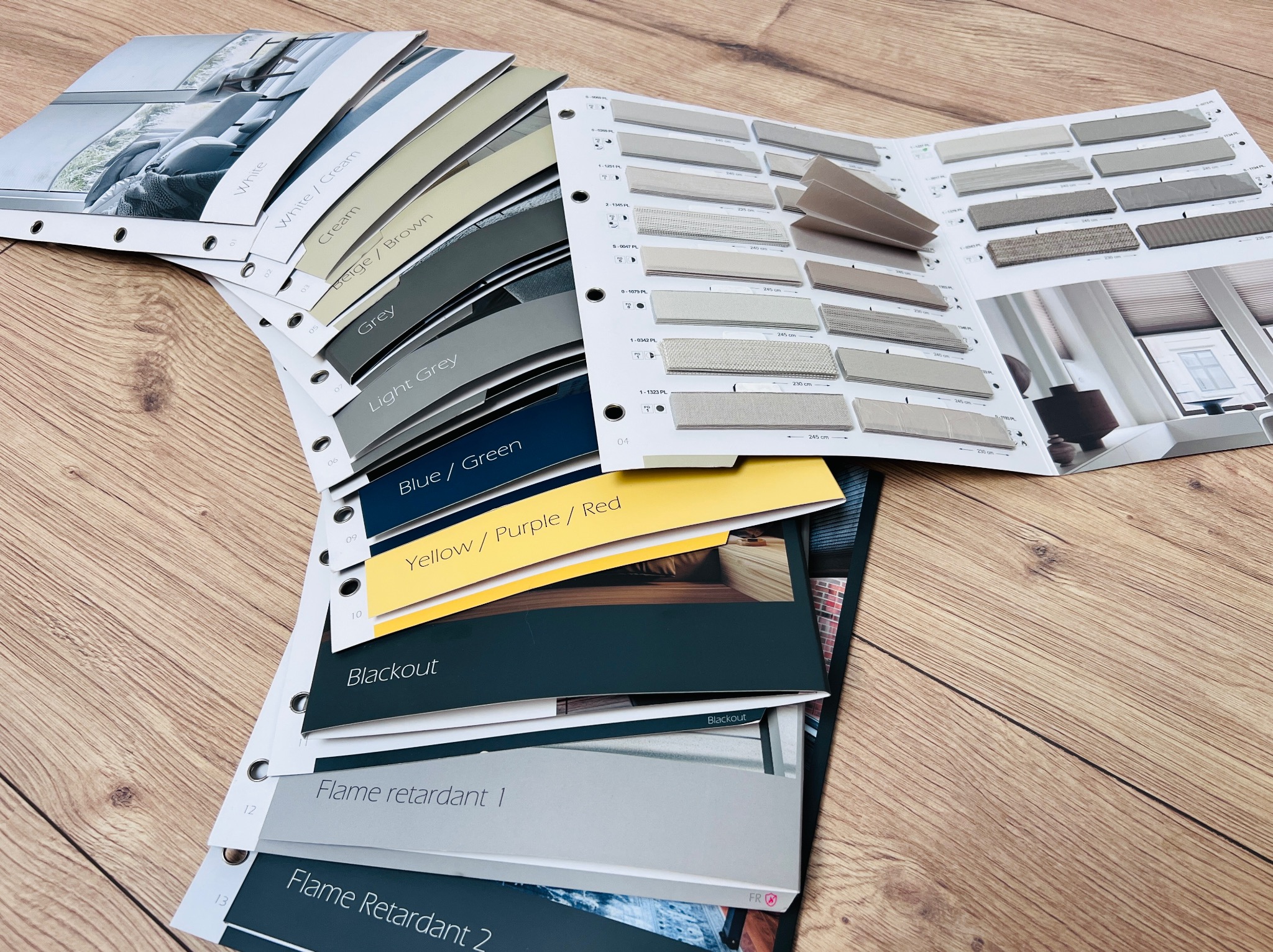 Katalog z próbkami tkanin i kolorów do rolet okiennych, w tym materiały zaciemniające i trudnopalne, ułożony na drewnianym blacie. Prezentacja wzornika.