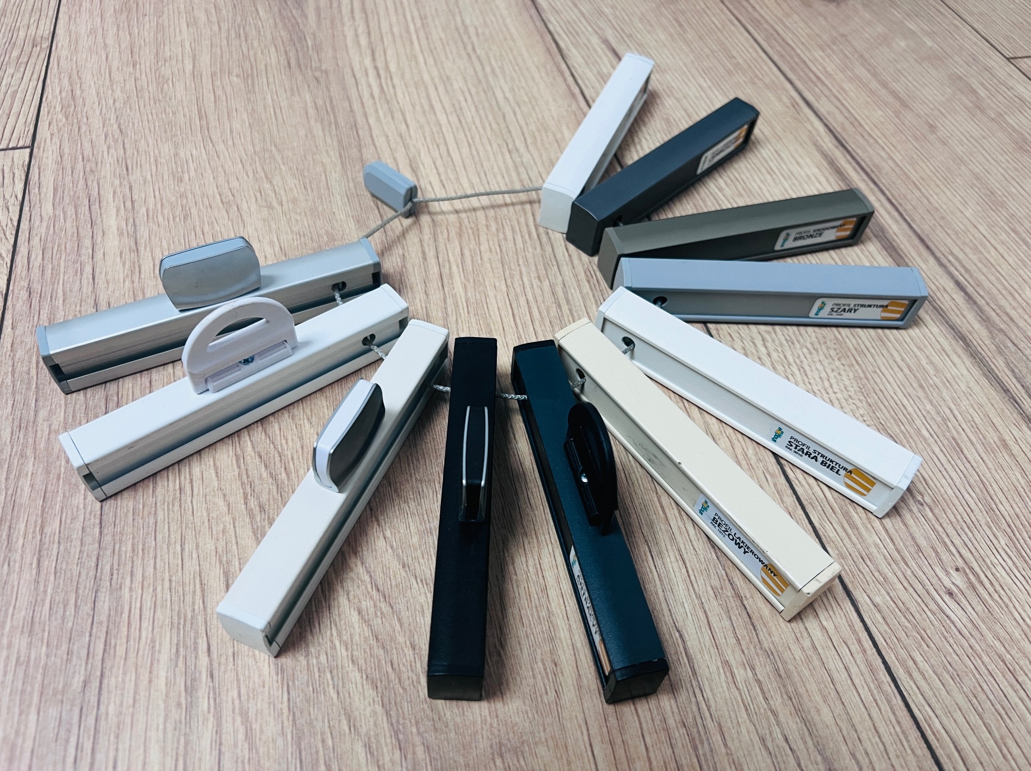 Przekrój profili do osłon okiennych w różnych kolorach (szary, biały, czarny, beżowy, brąz) ułożonych na drewnianym blacie. Produkcja i montaż w Pętkowicach.