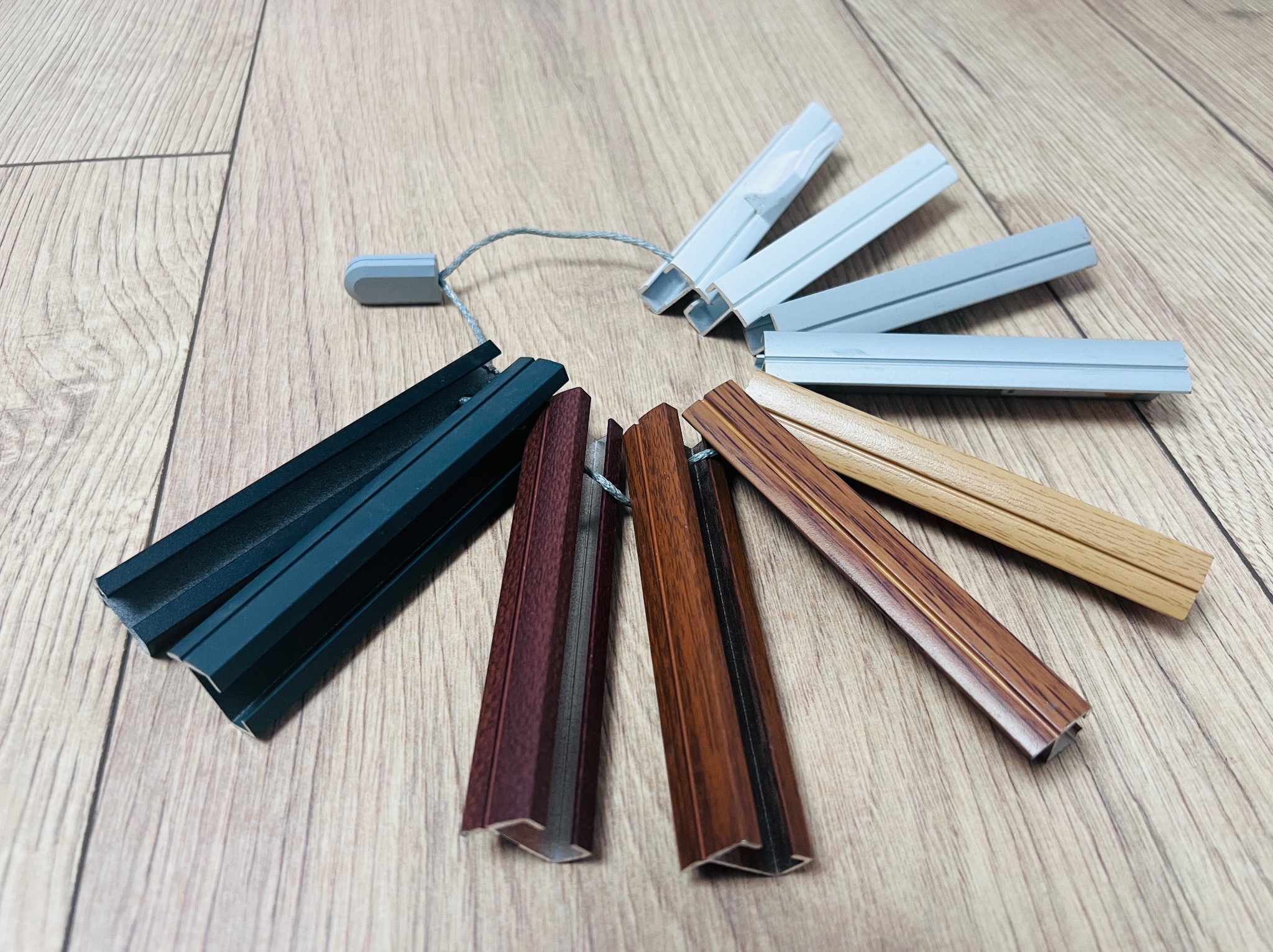Próbki kolorów i materiałów do żaluzji i rolet wewnętrznych, ułożone wachlarzowo na drewnianej podłodze. Prezentacja różnych odcieni i faktur.